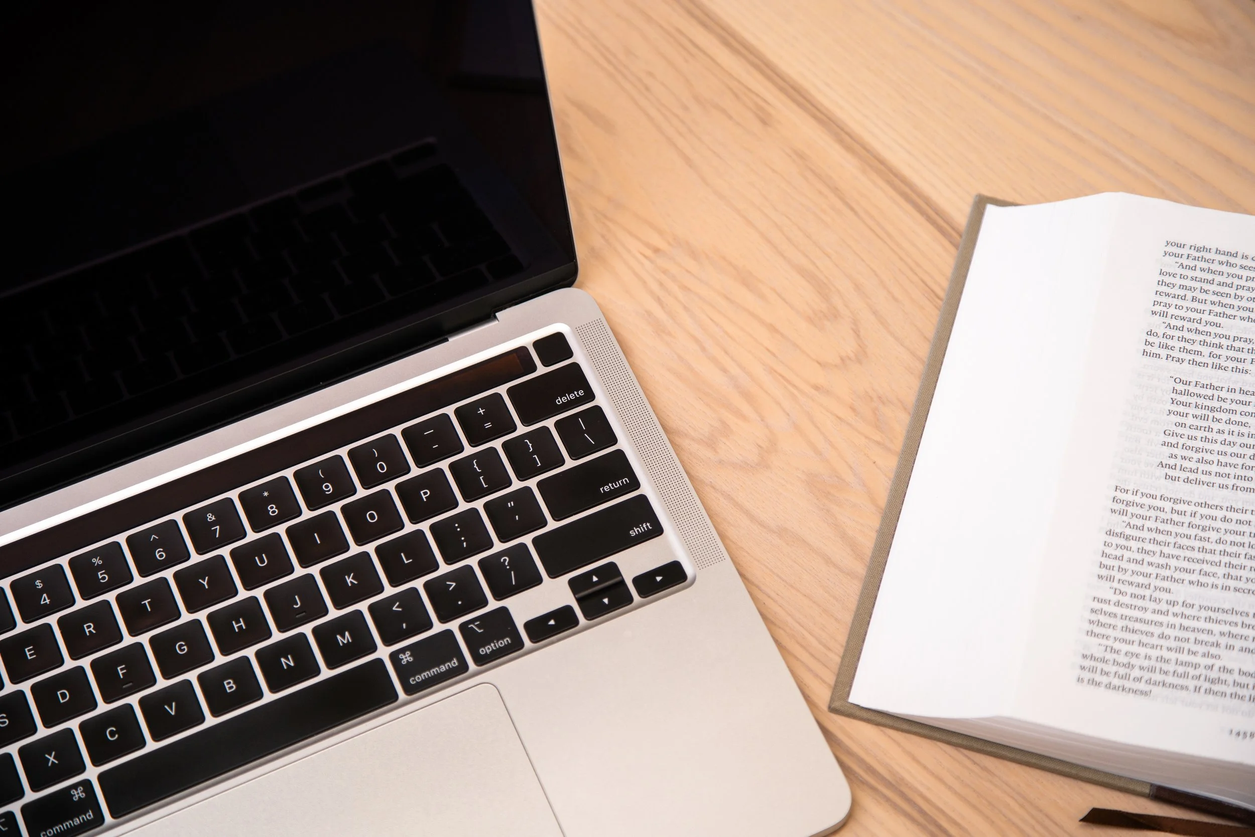 Part of a silver laptop with a black keyboard, open on a wooden desk next to a Bible with a brown cover and white pages.