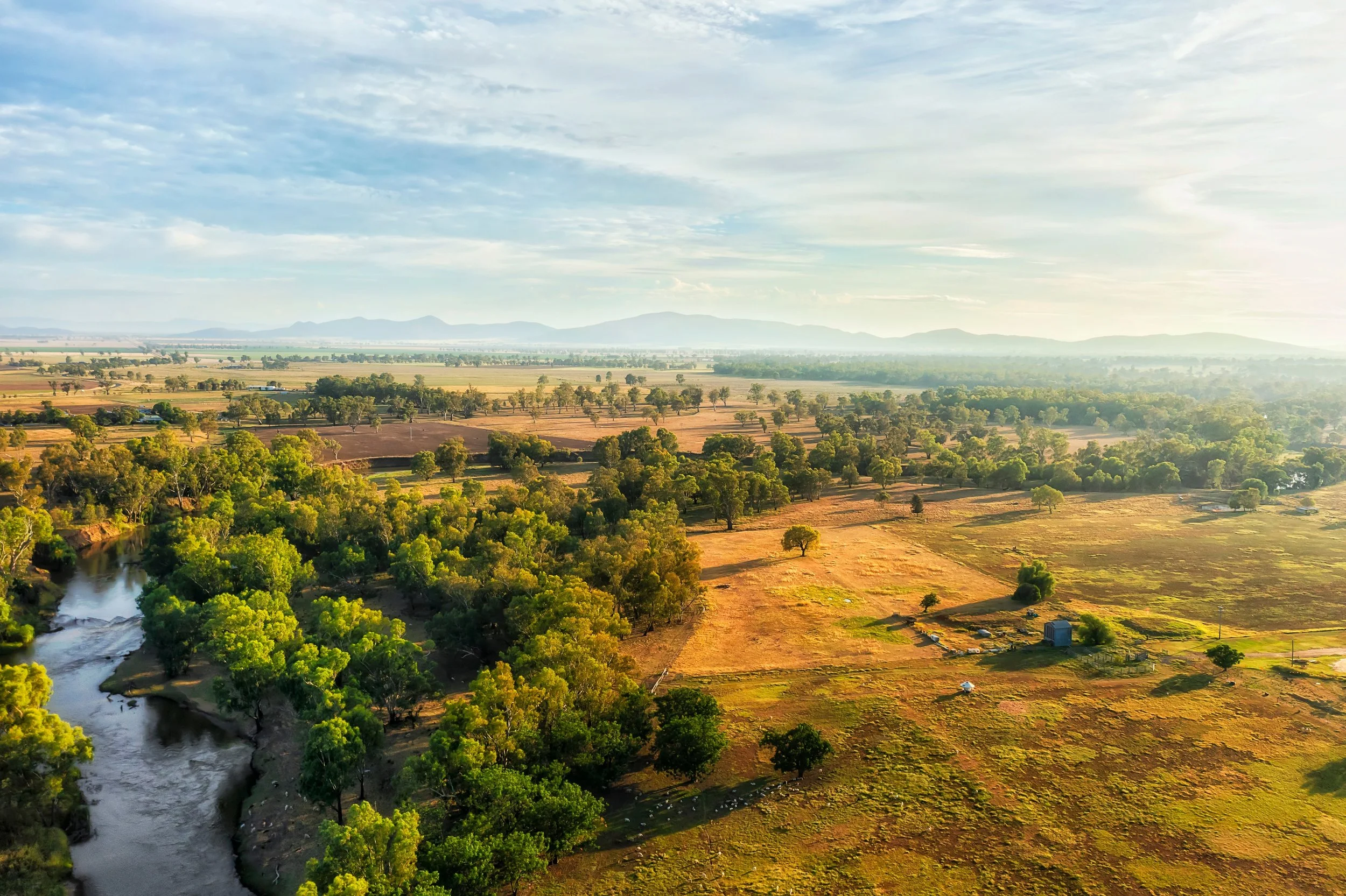Aerial view of a rural landscape featuring a winding river, green trees, open fields, and distant mountains under a partly cloudy sky.