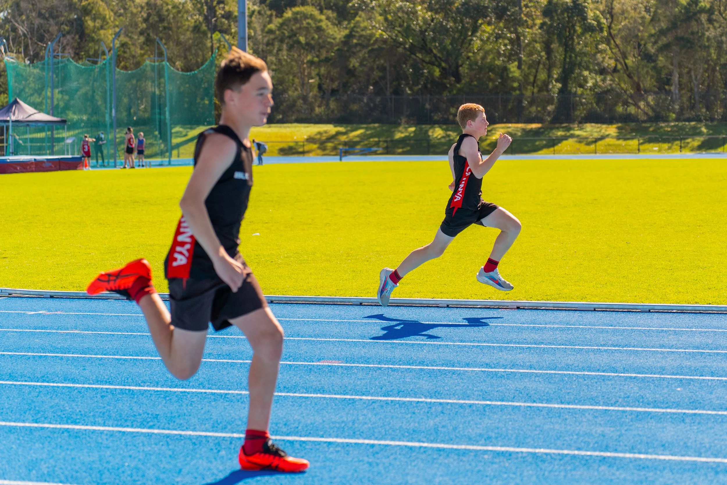 Two young male athletes in black running uniforms with red accents competing in a race on a blue track. The runner in the background is mid-air, while the other is closer to the camera, both showing determined focus.