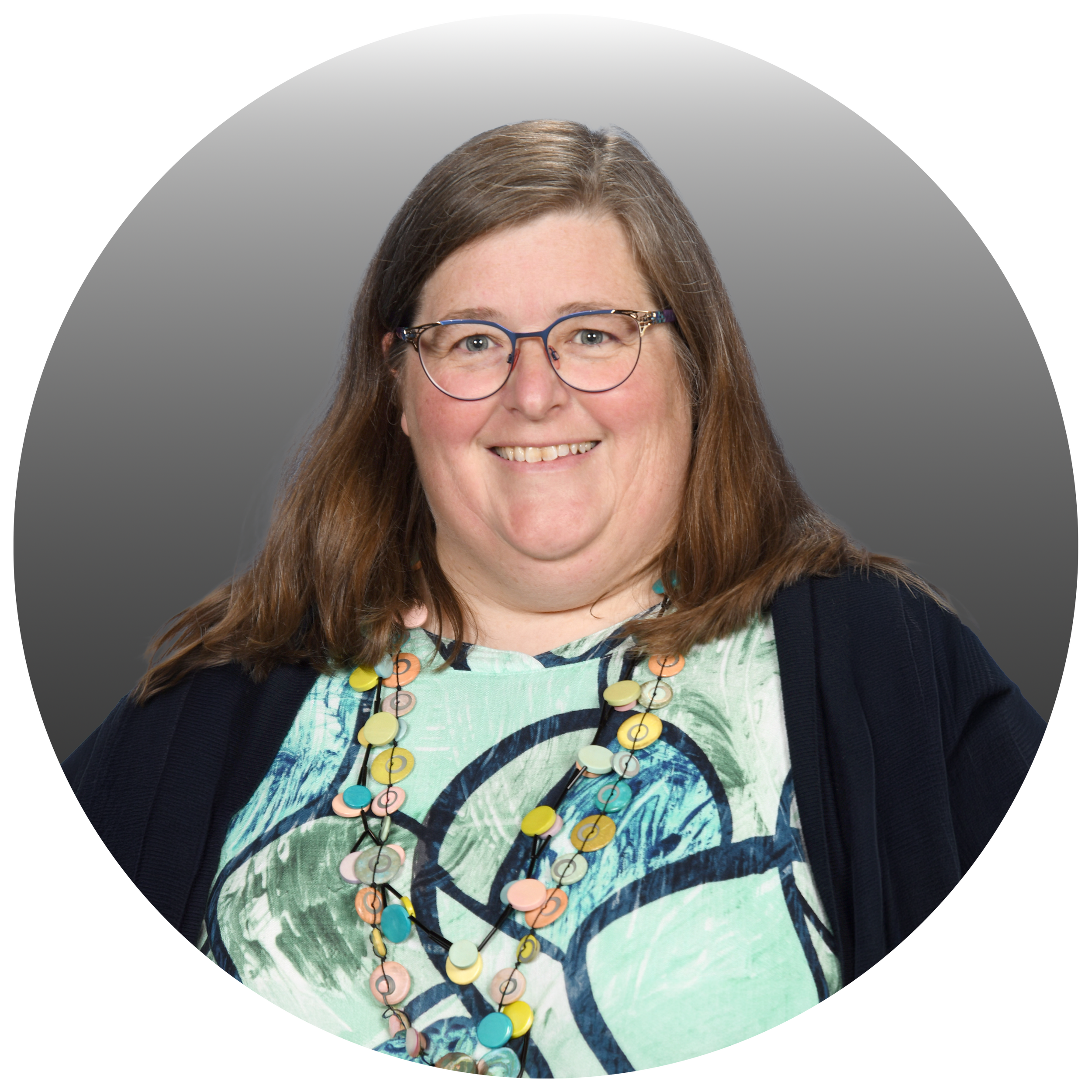 A woman with long brown hair, glasses, and a colorful patterned shirt smiling in front of a gray and black background.