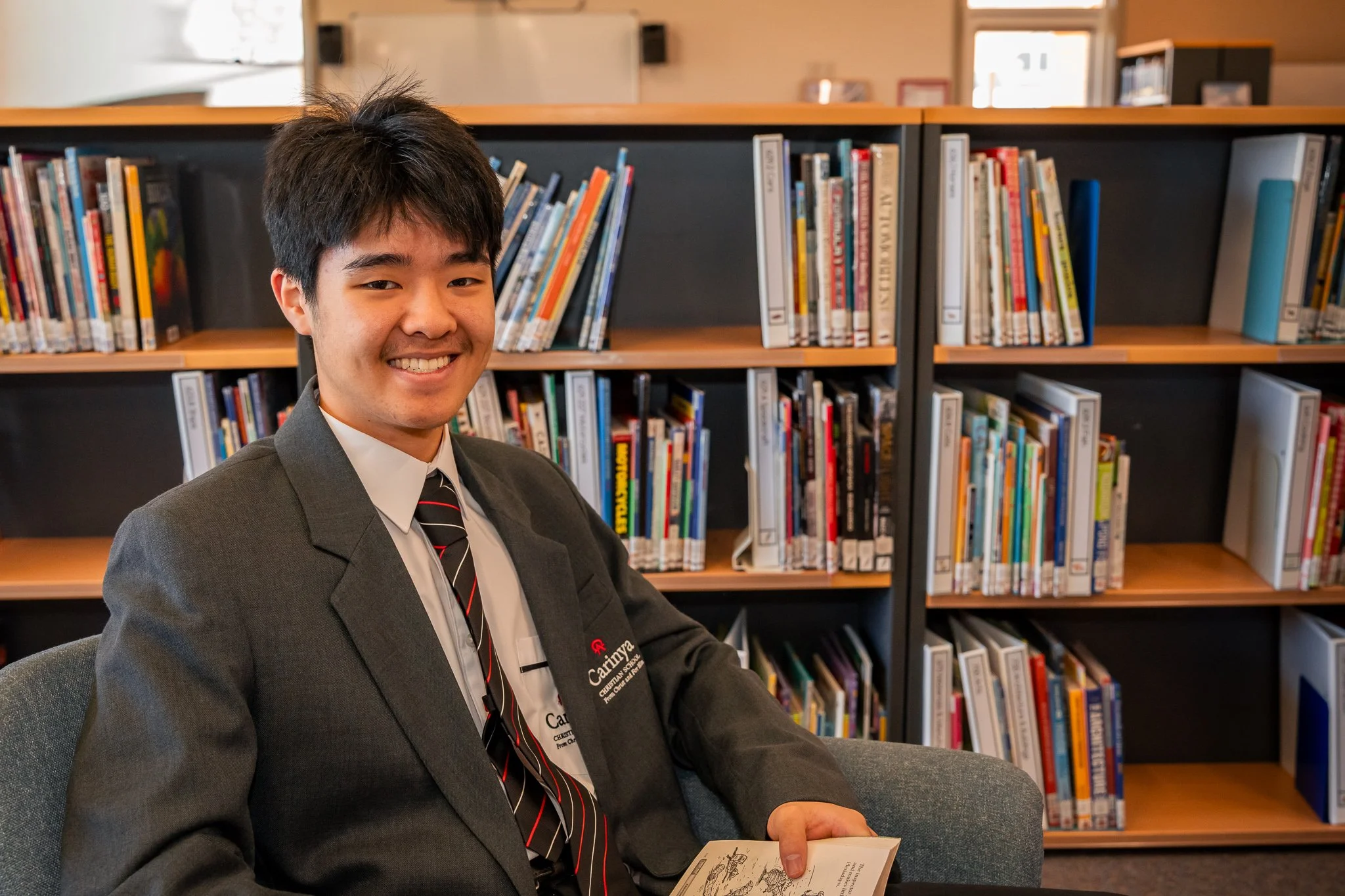 Young man in a dark suit and striped tie smiling while sitting in a library, holding a book with a bookshelf filled with books in the background.