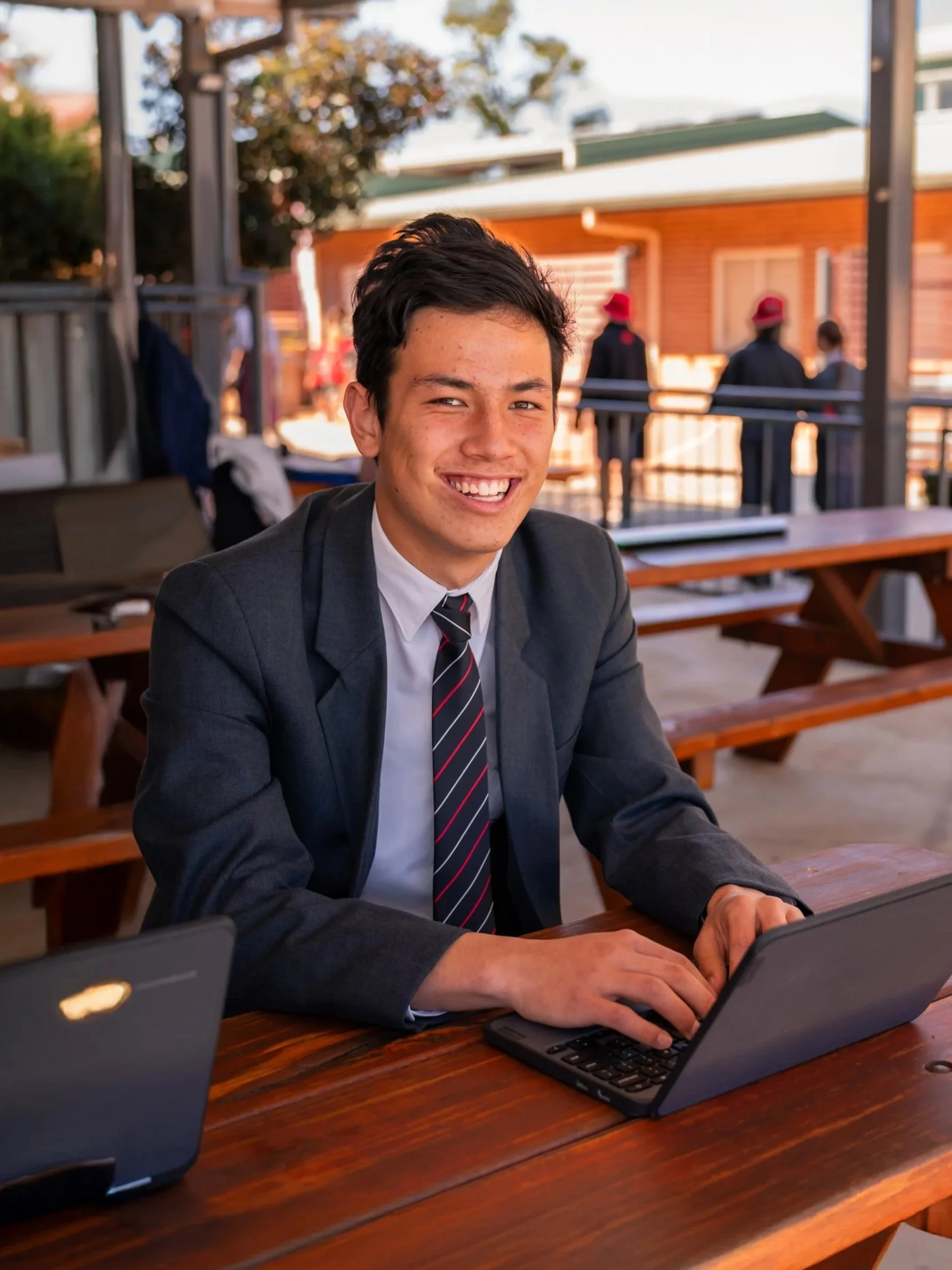 A young man in a business suit and tie sitting at an outdoor wooden picnic table, smiling at the camera with a laptop in front of him.