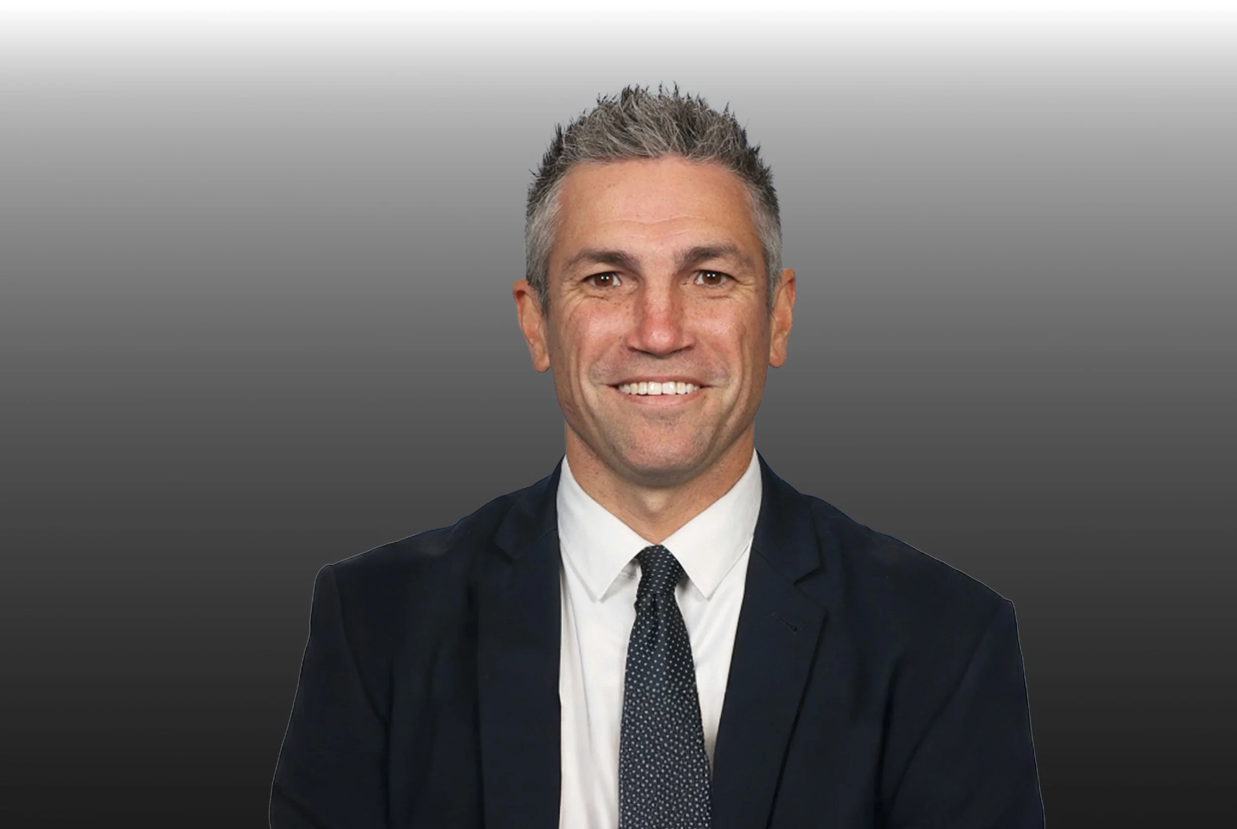 A professional headshot of a middle-aged man with short gray hair, wearing a dark suit, white shirt, and dotted tie, smiling against a gray gradient background.