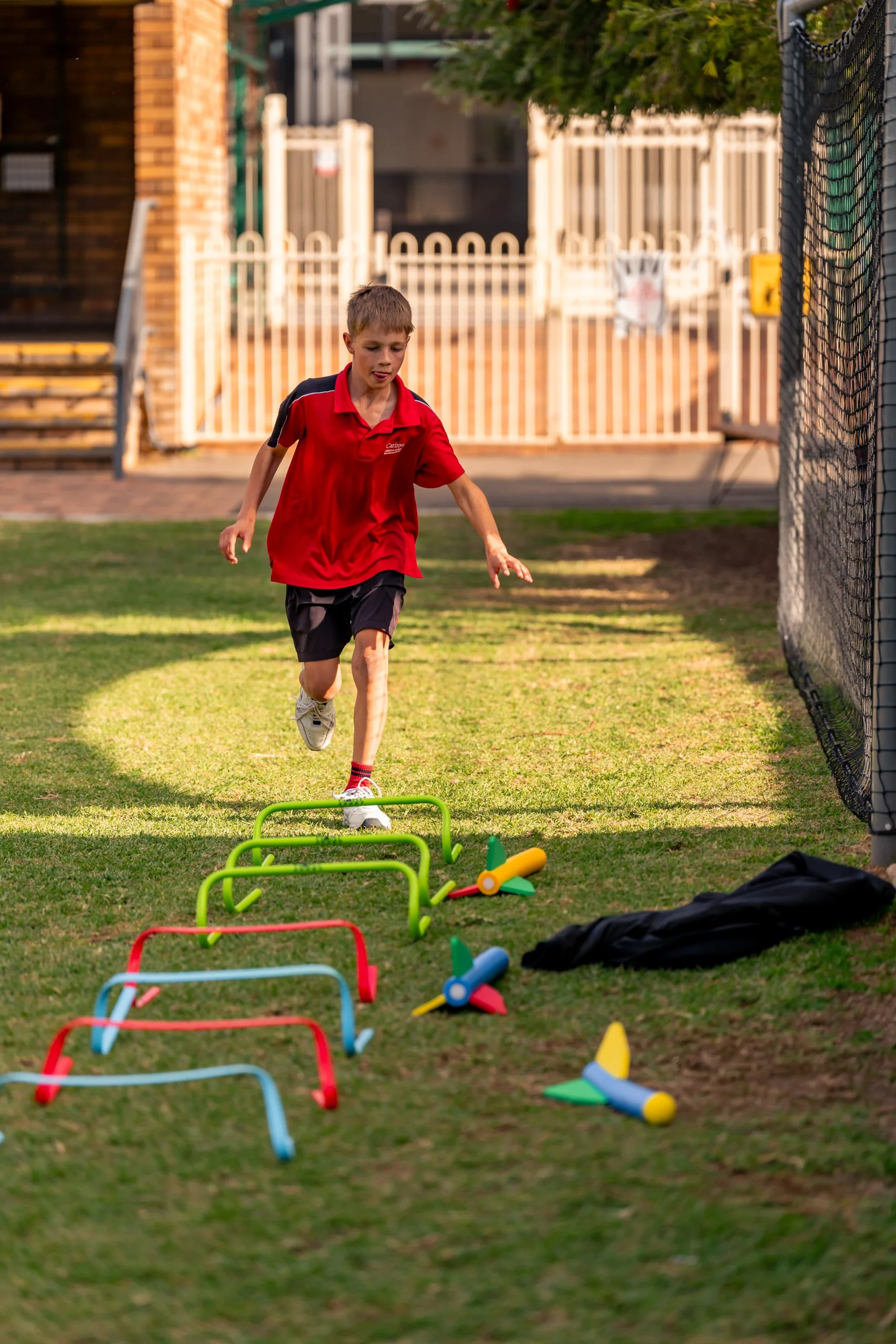 A young boy in a red shirt and black shorts running through an agility obstacle course outdoors on a grassy field, with hurdles and colorful cones.