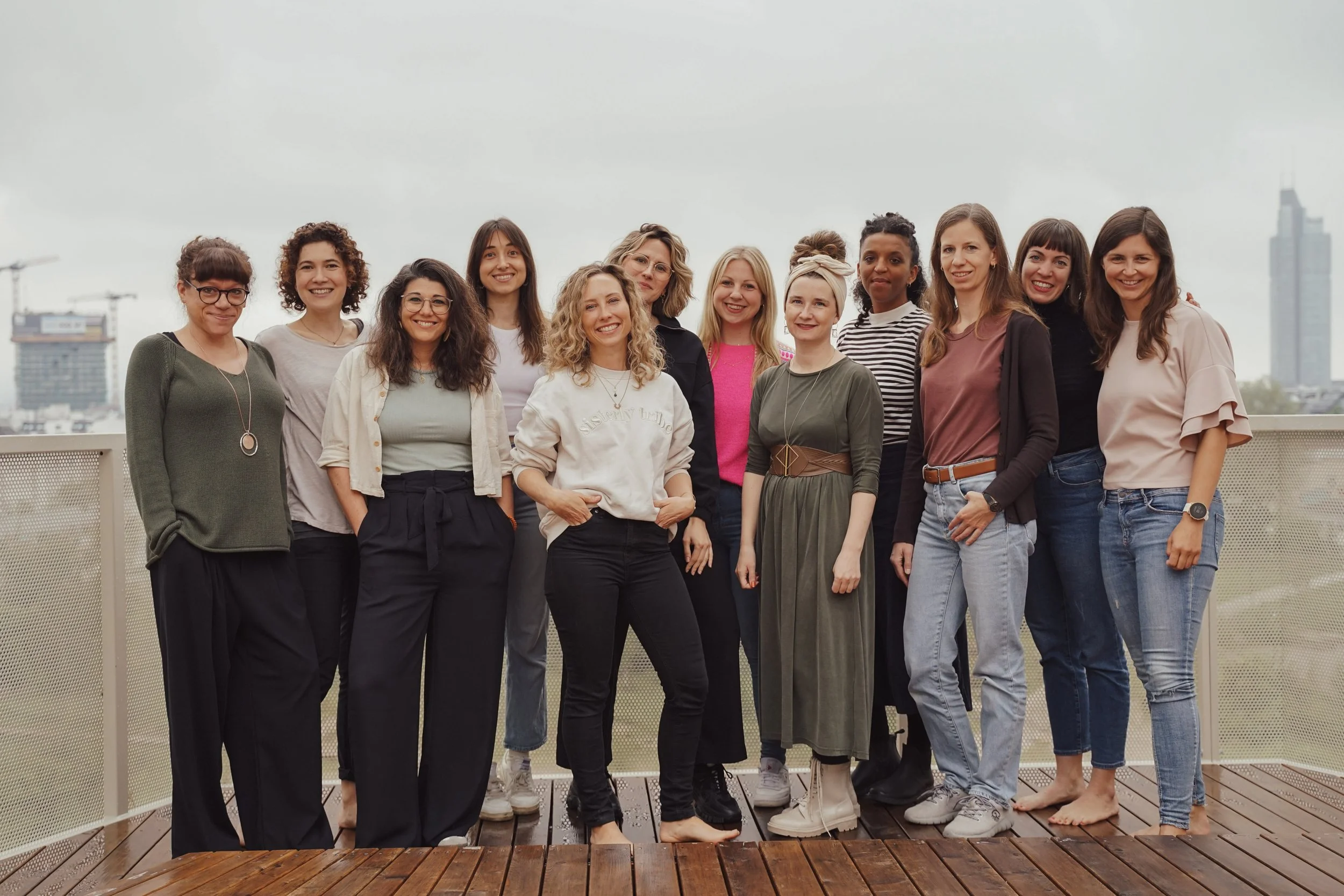 Eine Gruppe von zwölf Frauen steht auf einer Dachterrasse. Sie lächeln in die Kamera und tragen legere Kleidung. Im Hintergrund sind Baukräne und moderne Gebäude zu sehen.