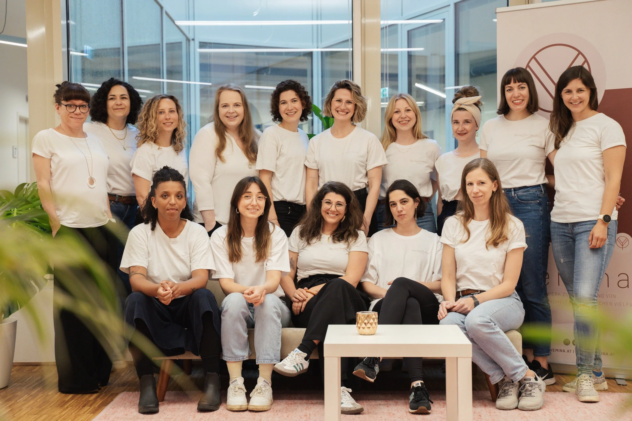 Eine Gruppe von 14 Frauen in weißen T-Shirts steht und sitzt zusammen in einem modernen Bürogebäude. Sie lächeln in die Kamera. Im Vordergrund ist ein Tisch mit einer Kerze zu sehen, im Hintergrund sind große Glasfenster.