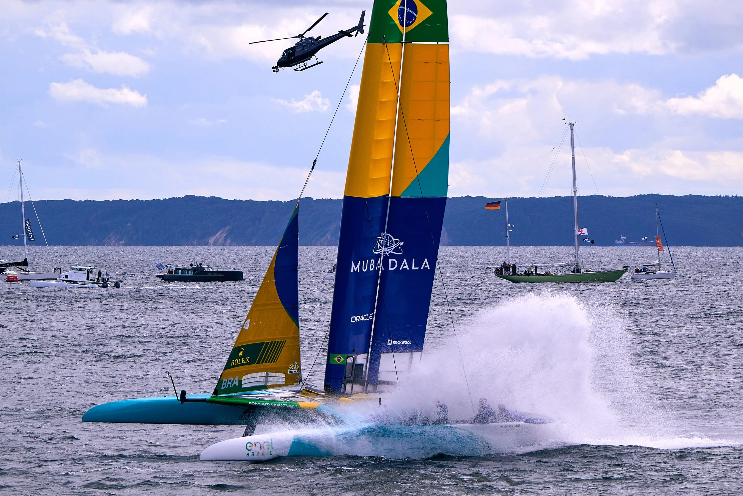A racing sailboat with brightly colored sails speeding across the water, with a helicopter flying overhead and several boats in the background on a cloudy day.