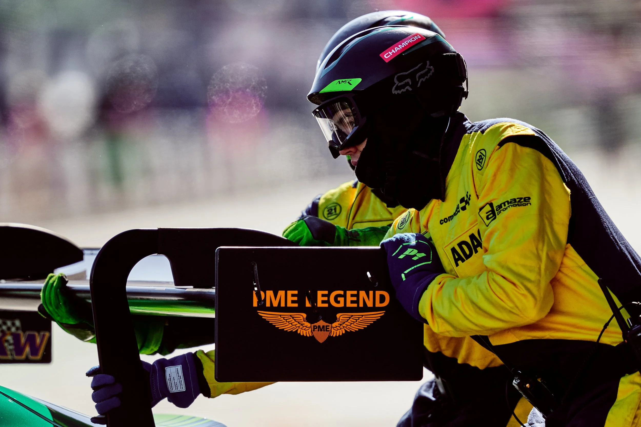 Person in a yellow safety jacket and black helmet working on a racing boat with a computer, held by another person wearing a blue and green outfit. The background is blurred and colorful.