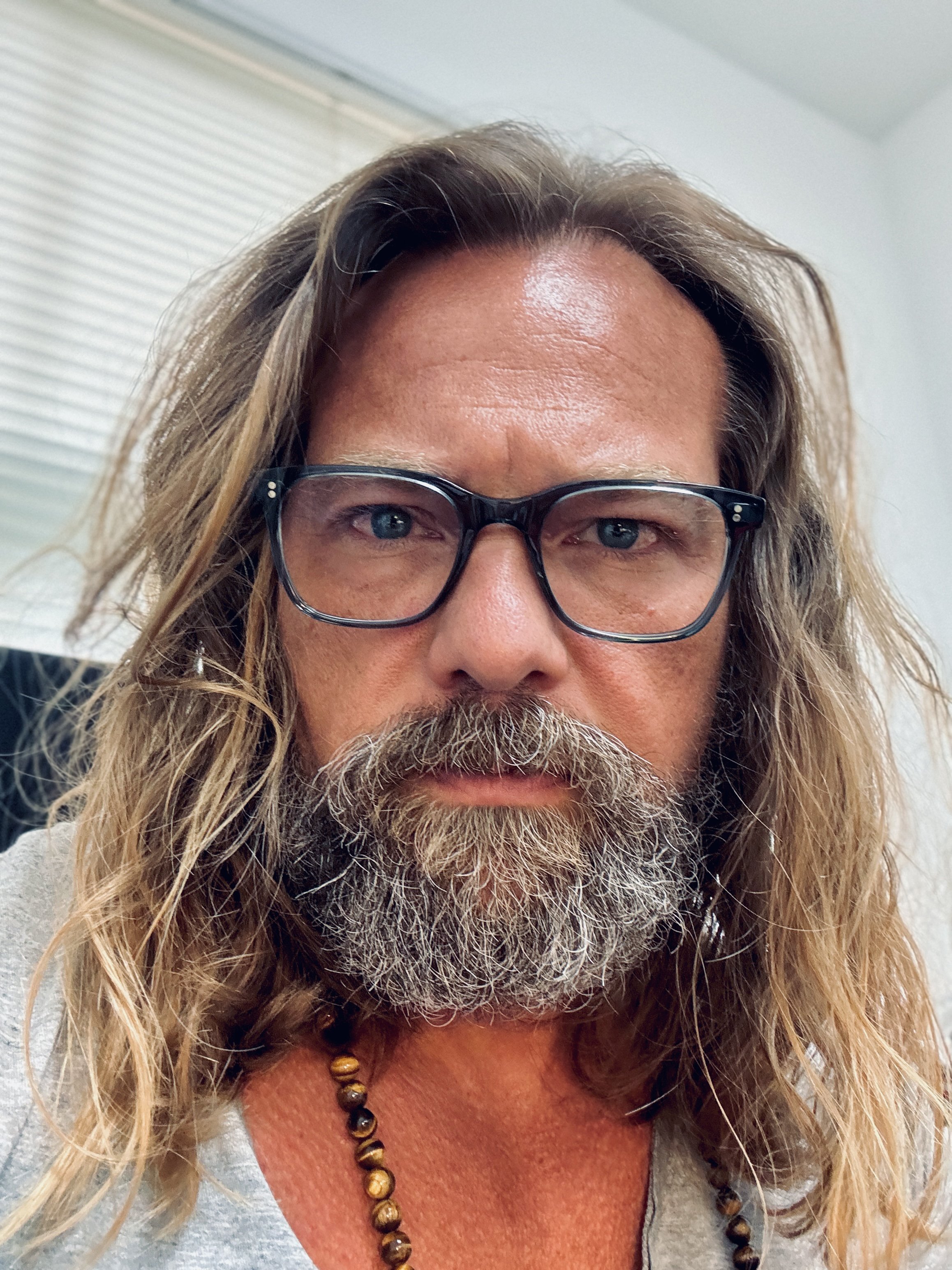 Close-up of a middle-aged man with long, wavy, blonde hair, gray beard, and glasses, wearing a gray shirt and a beaded necklace.