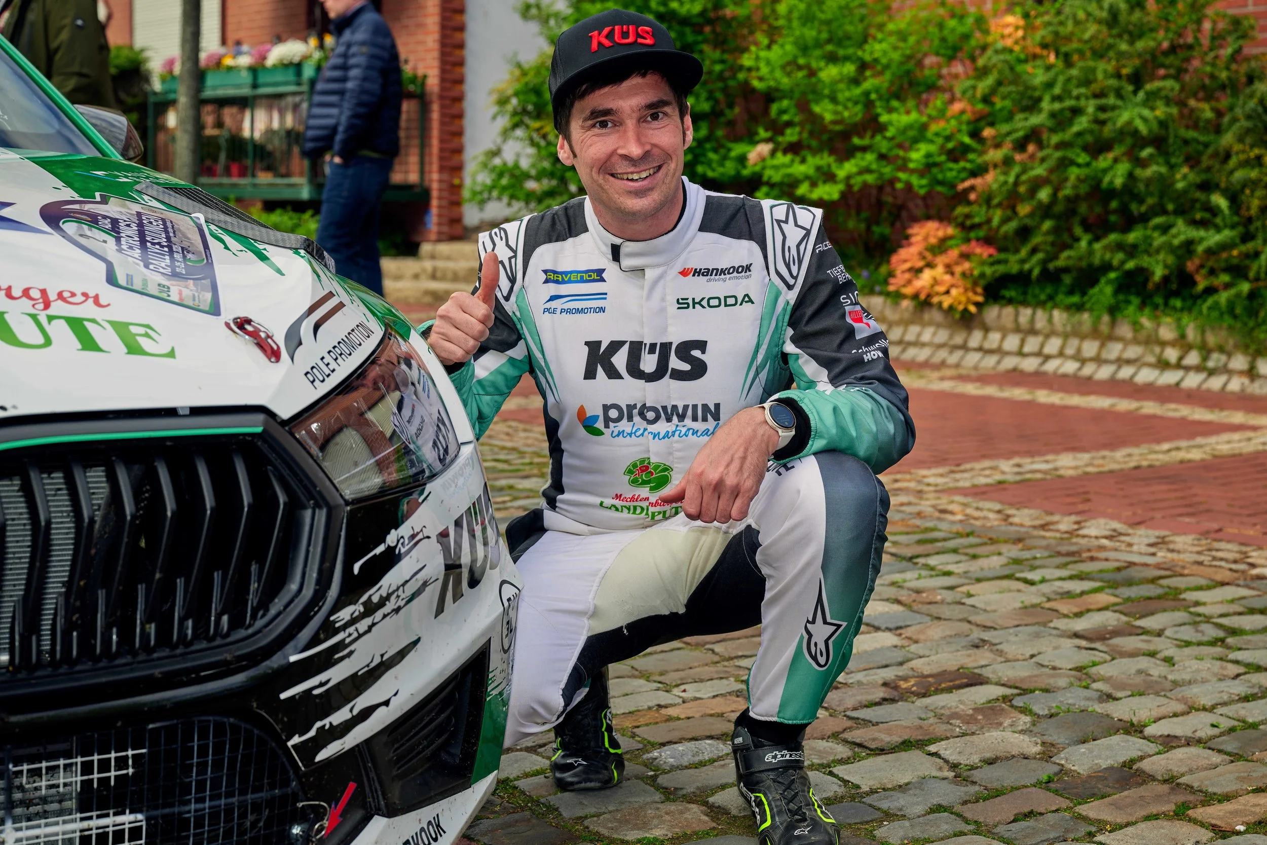 A man in a racing suit crouches next to a race car, smiling and giving a thumbs-up. He wears a black cap with red lettering, black and green racing shoes, and is outdoors on a brick-paved area with green foliage in the background.
