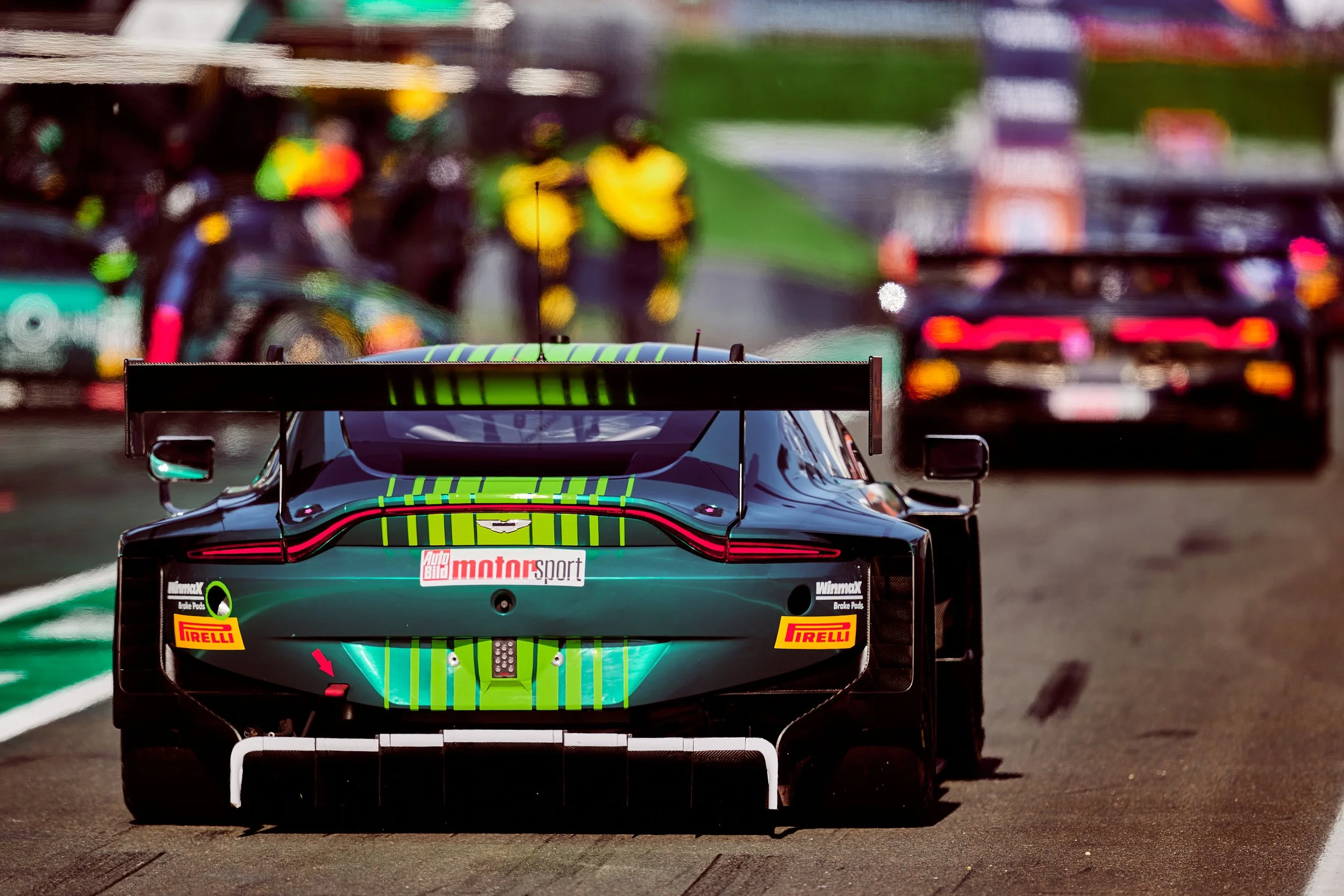 Rear view of a race car on the track, with a large rear wing and various sponsor stickers, amidst blurred background of other racing trucks and equipment.