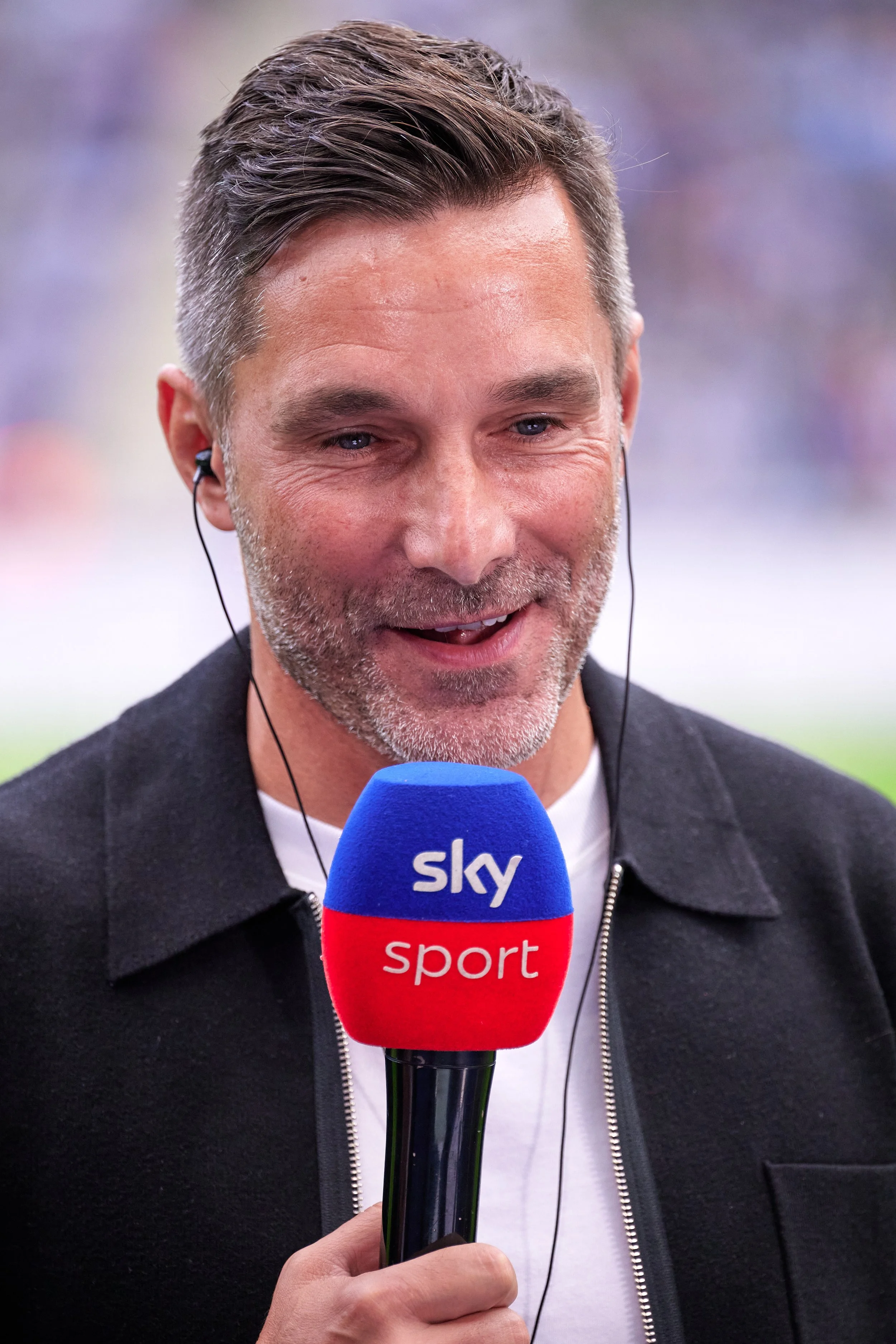 A man with short, dark hair and a beard is smiling while speaking into a microphone labeled "sky sport." He is wearing a black jacket with a visible white shirt underneath and appears to be at a sports event.