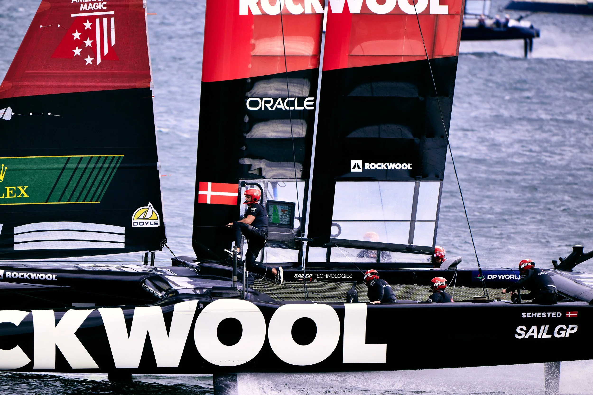 A racing sailboat with black and red sails featuring the logos 'Rockwool,' 'Oracle,' and 'Sehested.' The sailboat has a crew member sitting near the helm and others in black gear on the deck. The background shows water with a small boat and distant shoreline.
