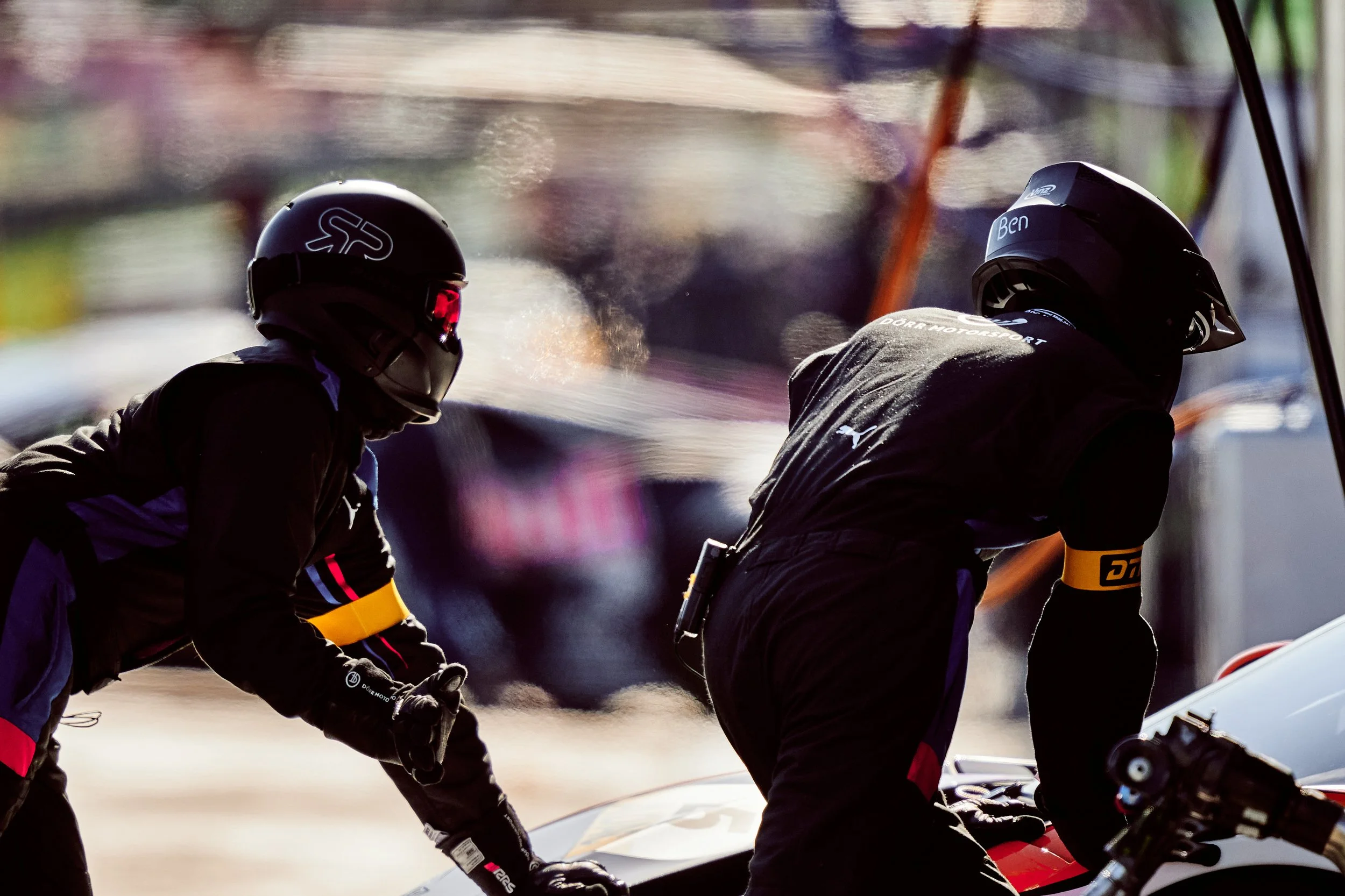 Two race car drivers wearing helmets and racing gear working on a race car during a race or practice session.