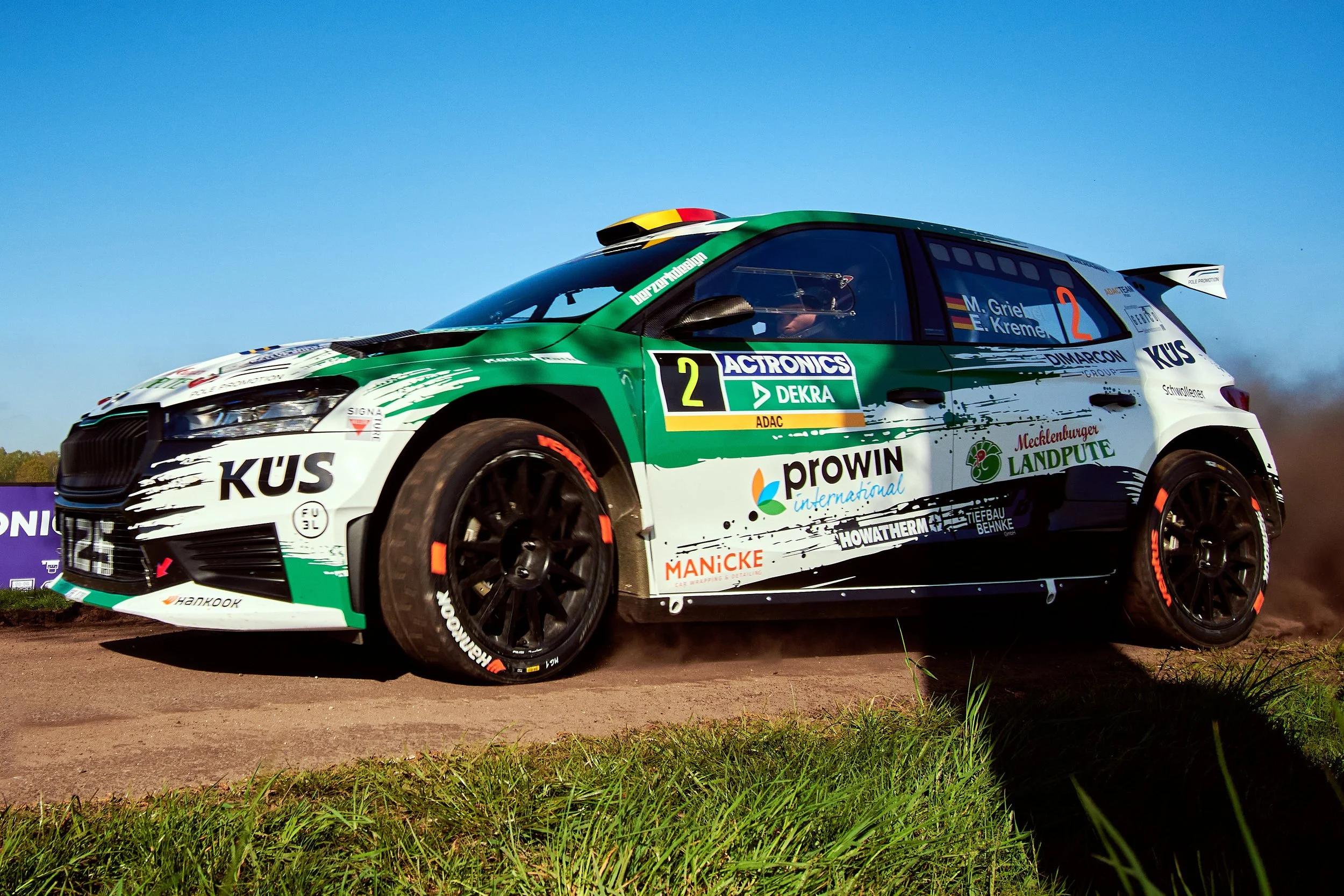 A rally car on dirt ground during a race, with a blue sky in the background. The car is green and white with various sponsor logos and the number 2 displayed on the side.