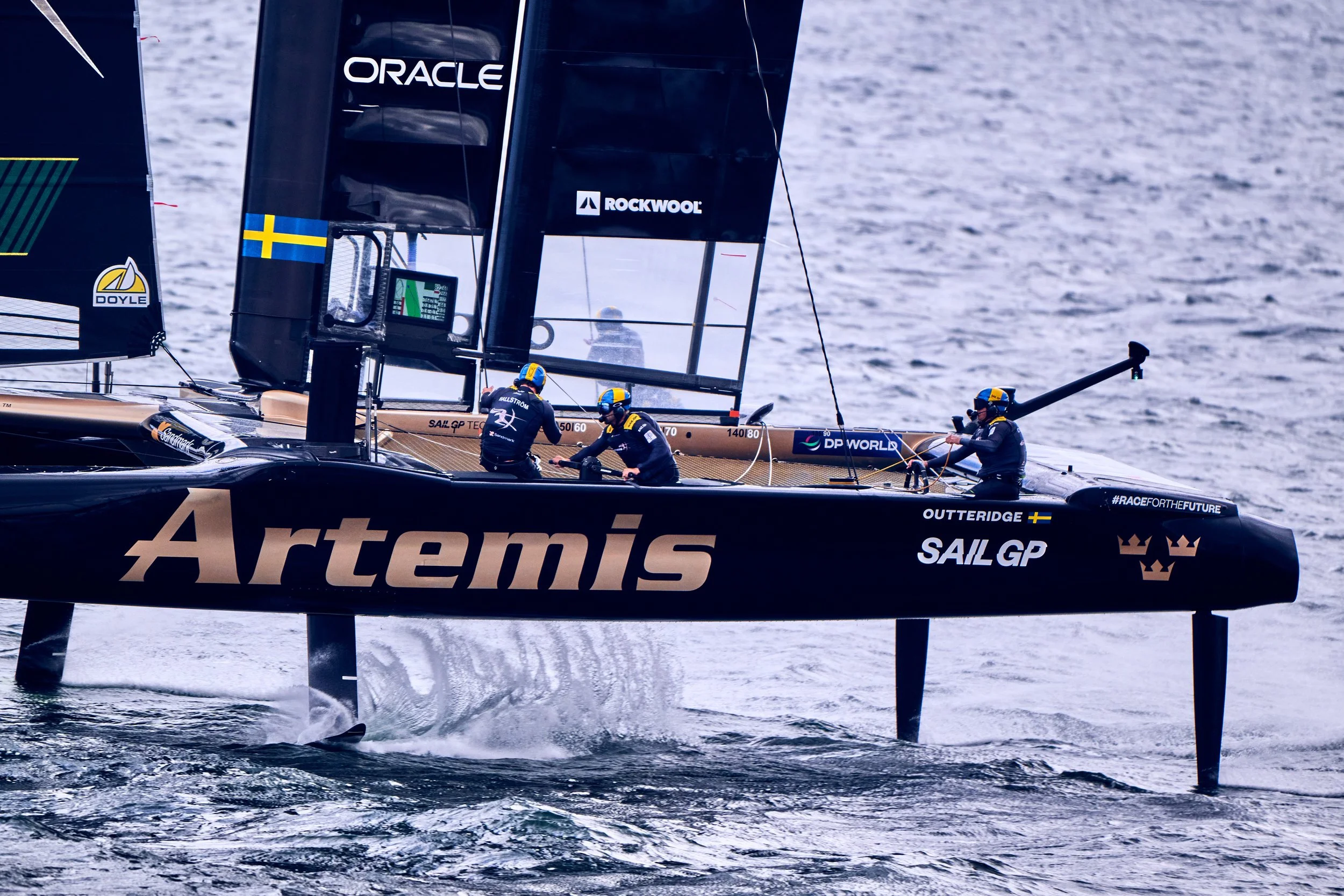 A high-performance racing sailboat on the water with crew members working on deck, displaying sponsorship logos including Oracle, Rockwool, and Artemis, during a competitive sailing event.