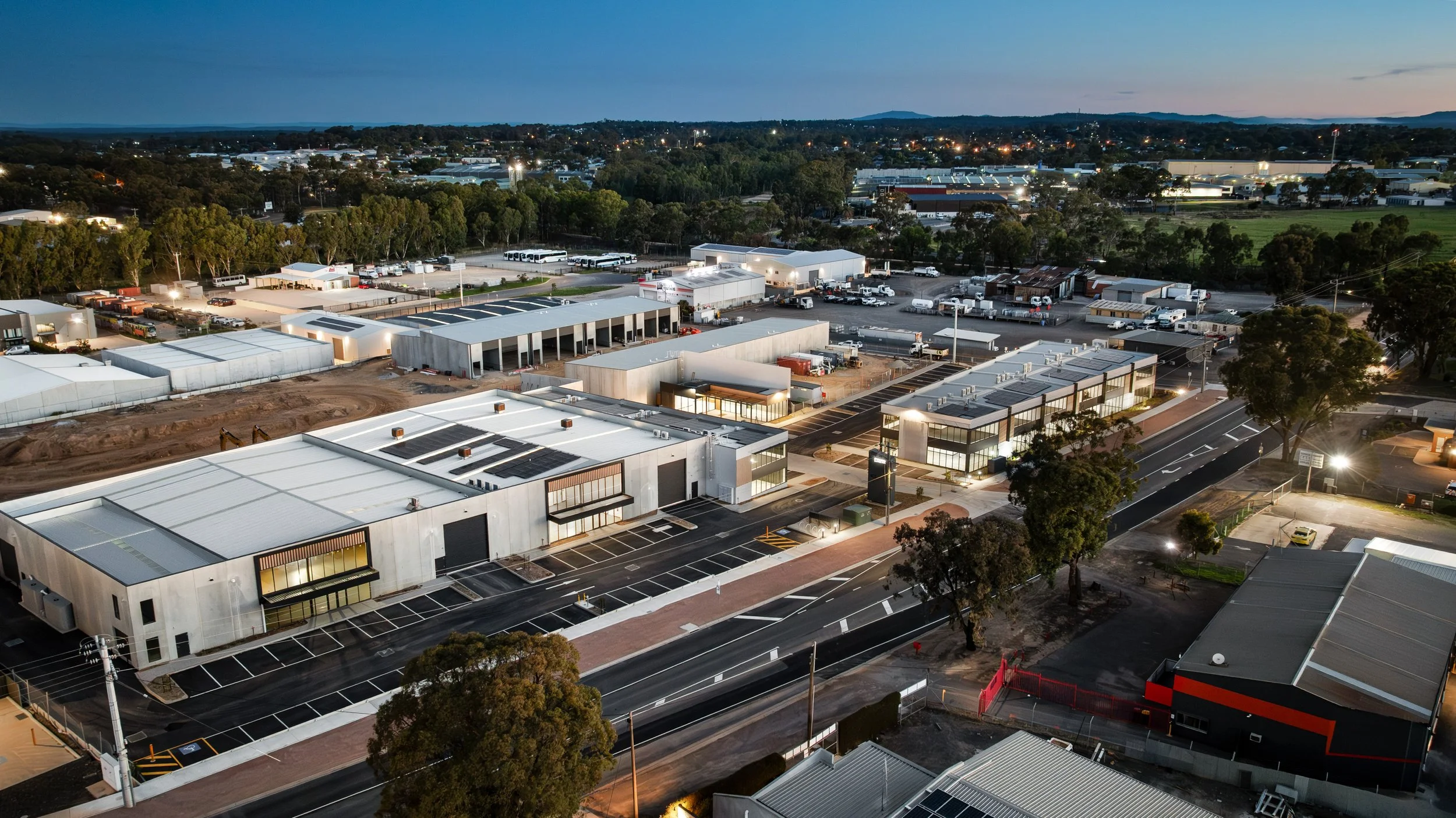 An aerial view of a commercial area at dusk with modern buildings, parking lots, and trees, with a cityscape and hills in the background.