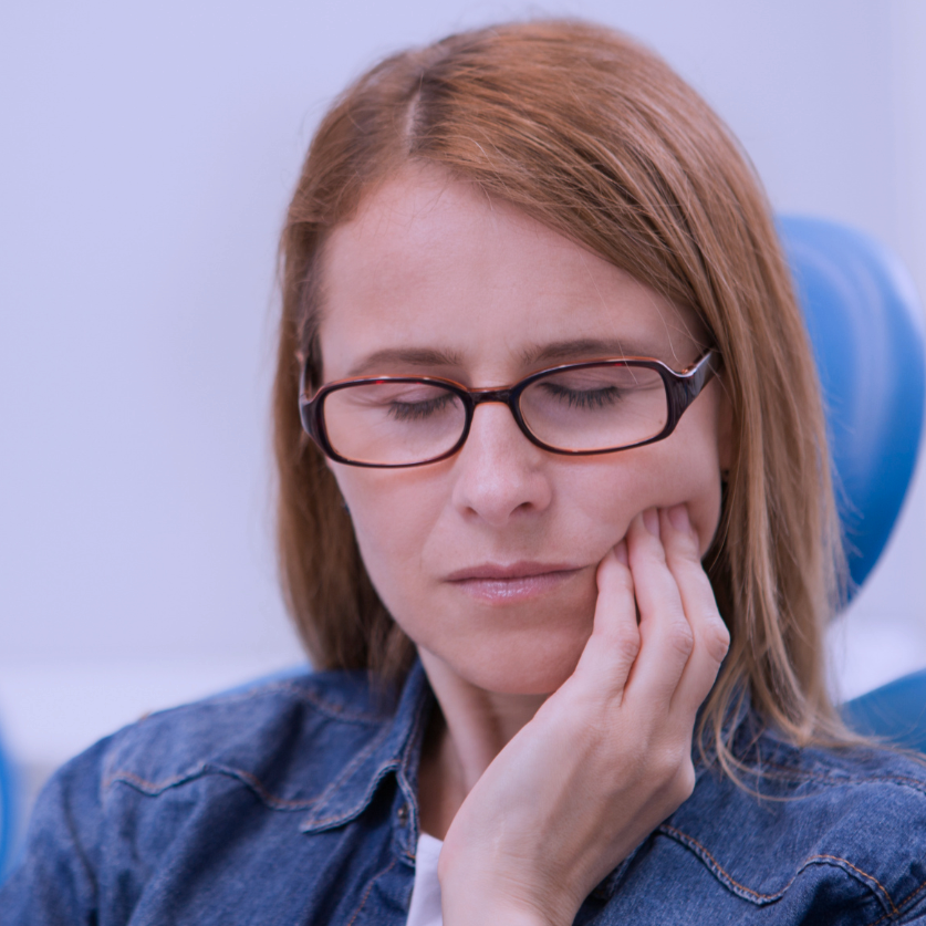 A woman with red hair and glasses sitting with her eyes closed, touching her cheek with her hand because of wisdom tooth pain.