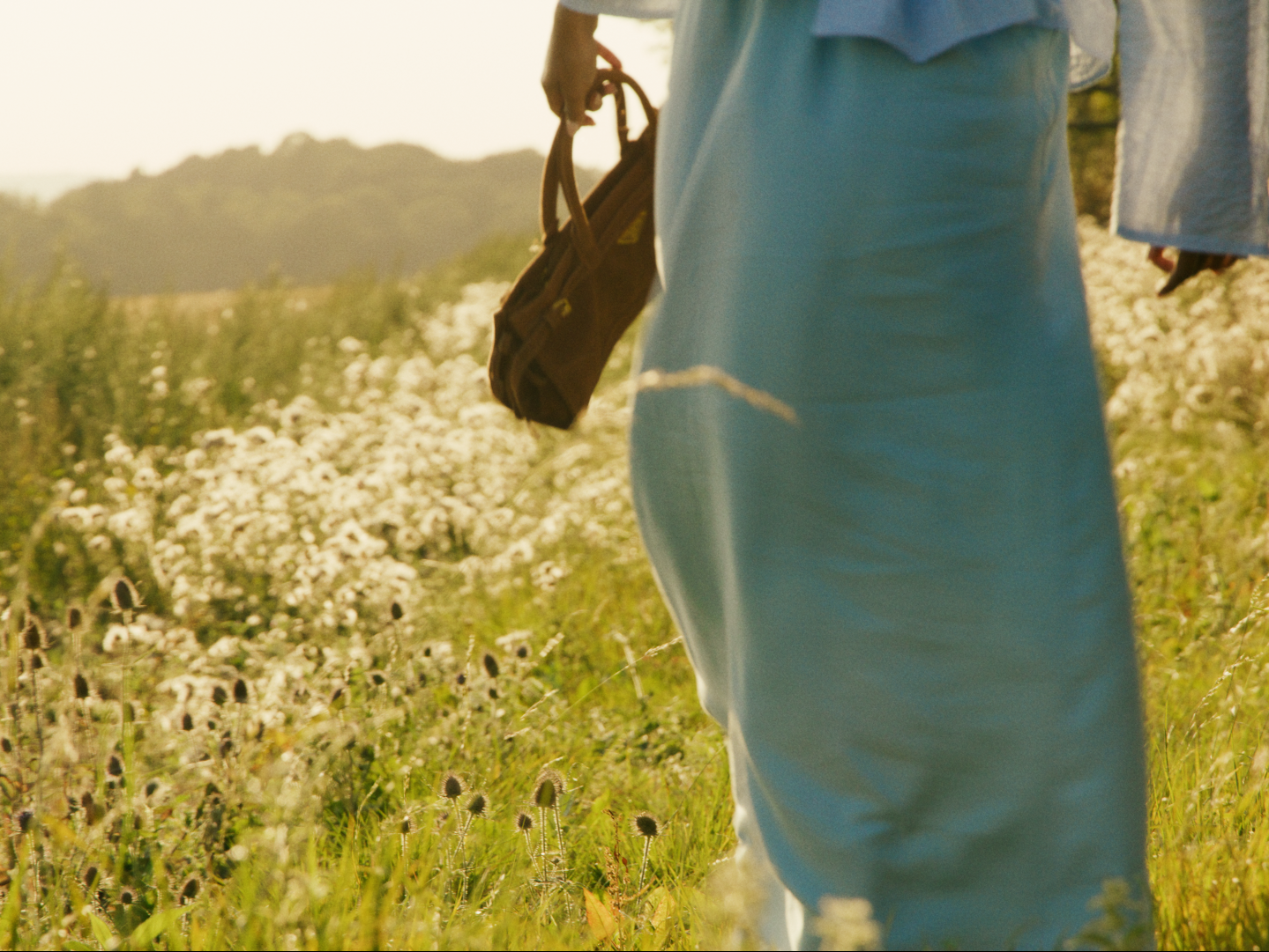 Close-up of a person in light blue pants holding a brown bag, standing in a field of white flowers with a mountain in the background.
