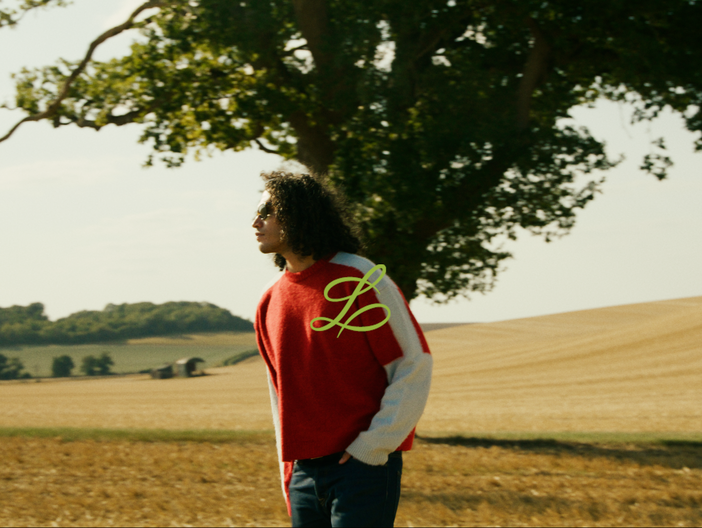 A woman standing outdoors in front of a large tree, wearing sunglasses and a red and white jacket, with a scenic, rural landscape in the background.