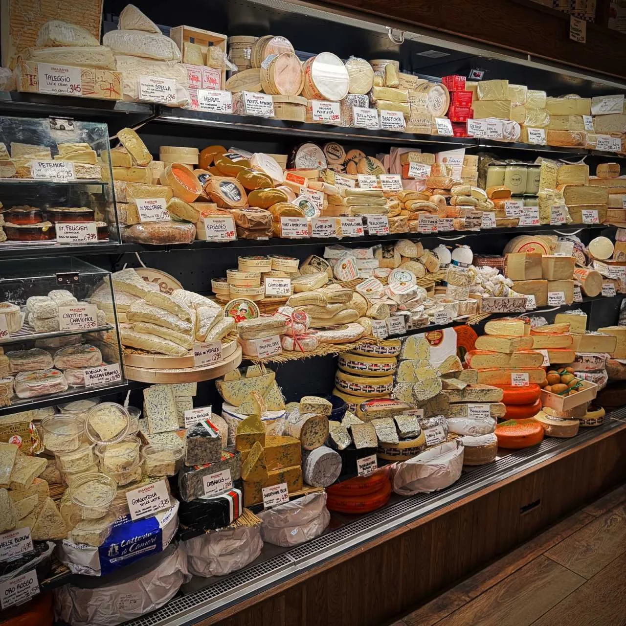 Ein Kühlregal im Shop De Kaaskamer in Amsterdam ist mit vielen Käsesorten wie Brie und Gorgonzola gefüllt. Preisschilder stecken in den verschiedenen Laiben auf den schwarzen Regalböden.