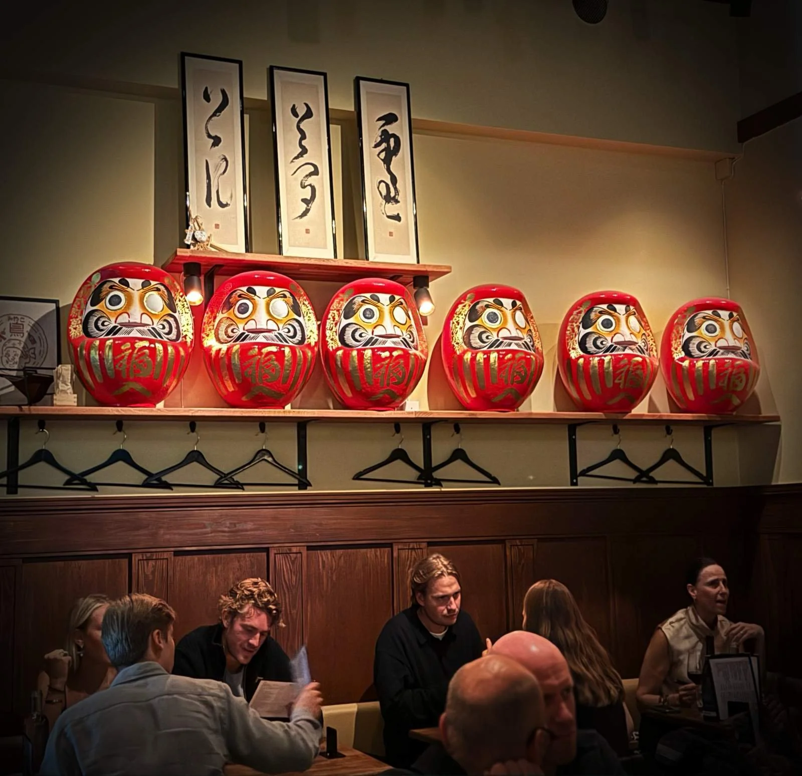Red Daruma figures on a shelf at Miyakodori restaurant in Stockholm with guests at tables and Japanese calligraphy on the wall.