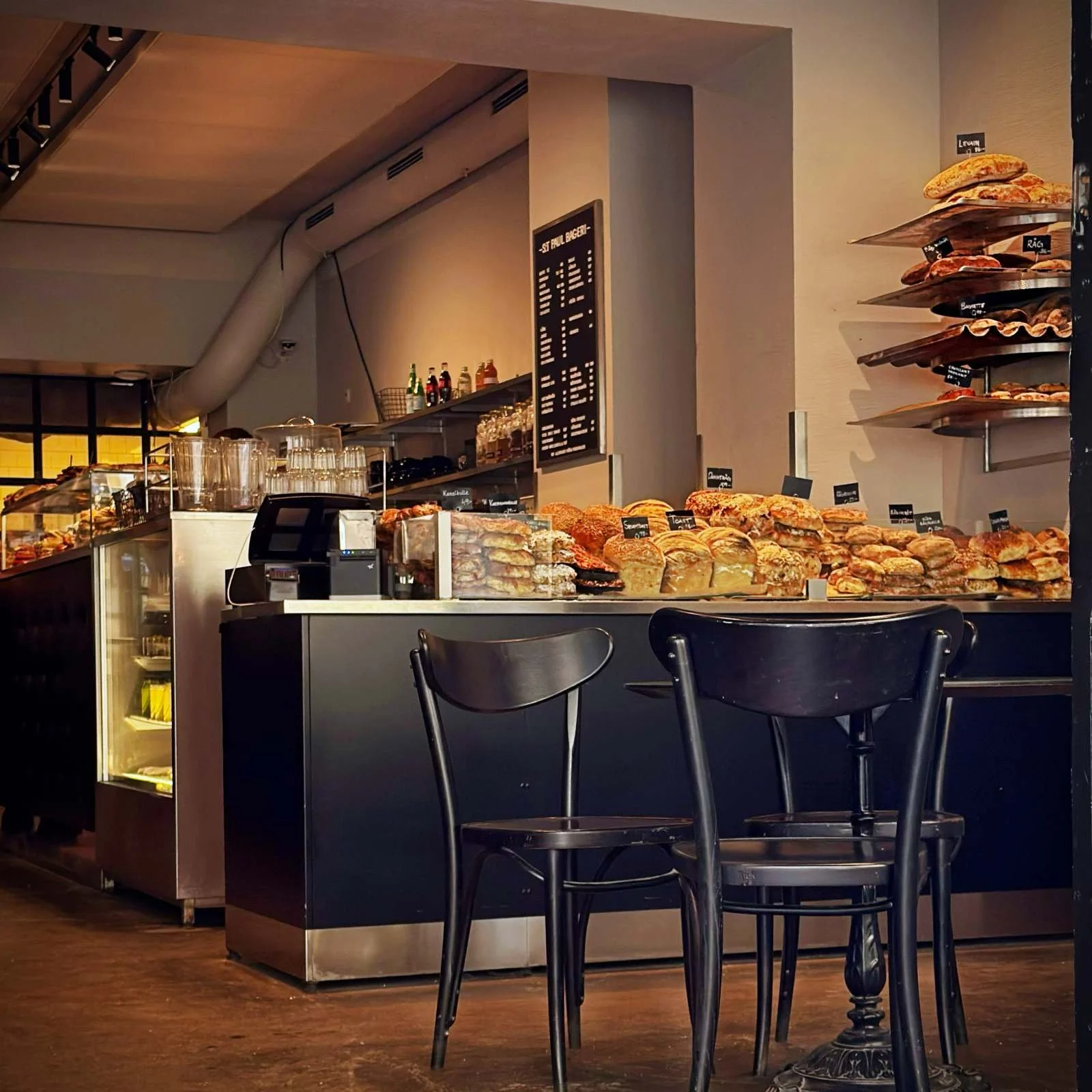A dark bakery counter stacked with a large selection of fresh breads and pastries. Two black wooden chairs stand in the foreground, and a price board hangs on the wall in the background.