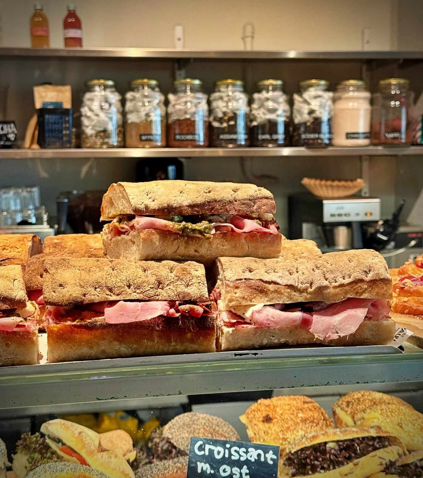 A stack of freshly prepared focaccia sandwiches filled with ham and pesto inside a display case. Spice jars and storage containers sit on a stainless steel shelf in the background.