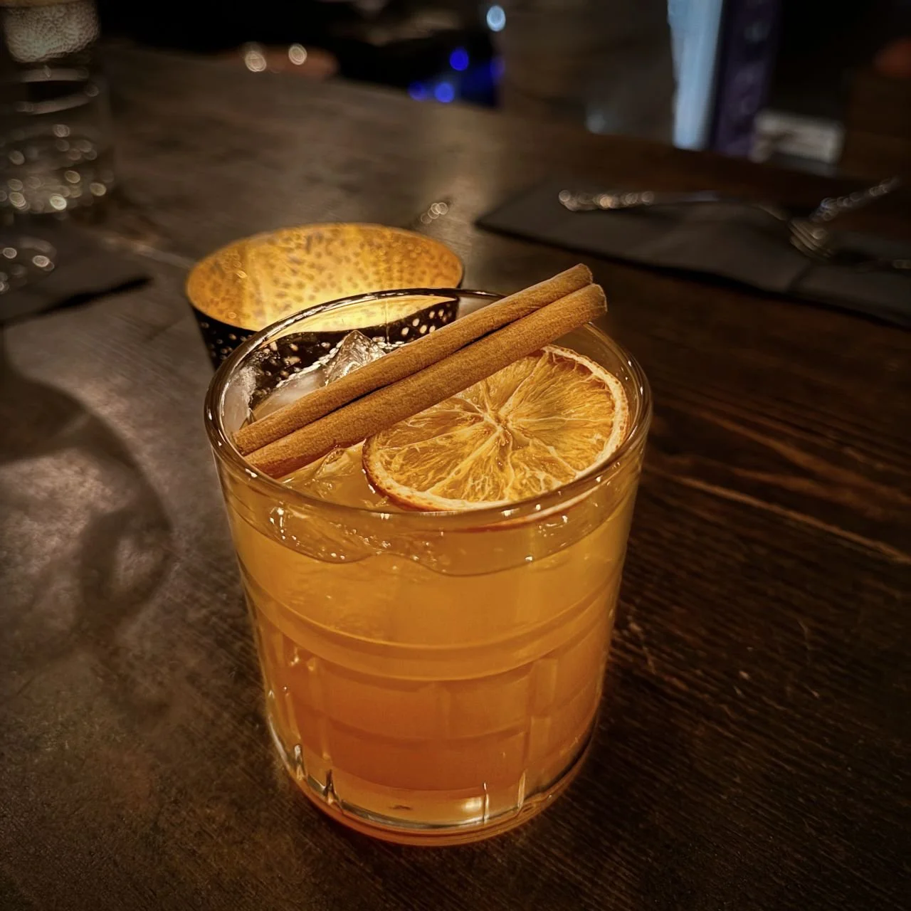 An orange cocktail with two cinnamon sticks and a dried orange slice at Night Kitchen restaurant in Berlin. The glass sits on a dark wooden table next to a lit candle.