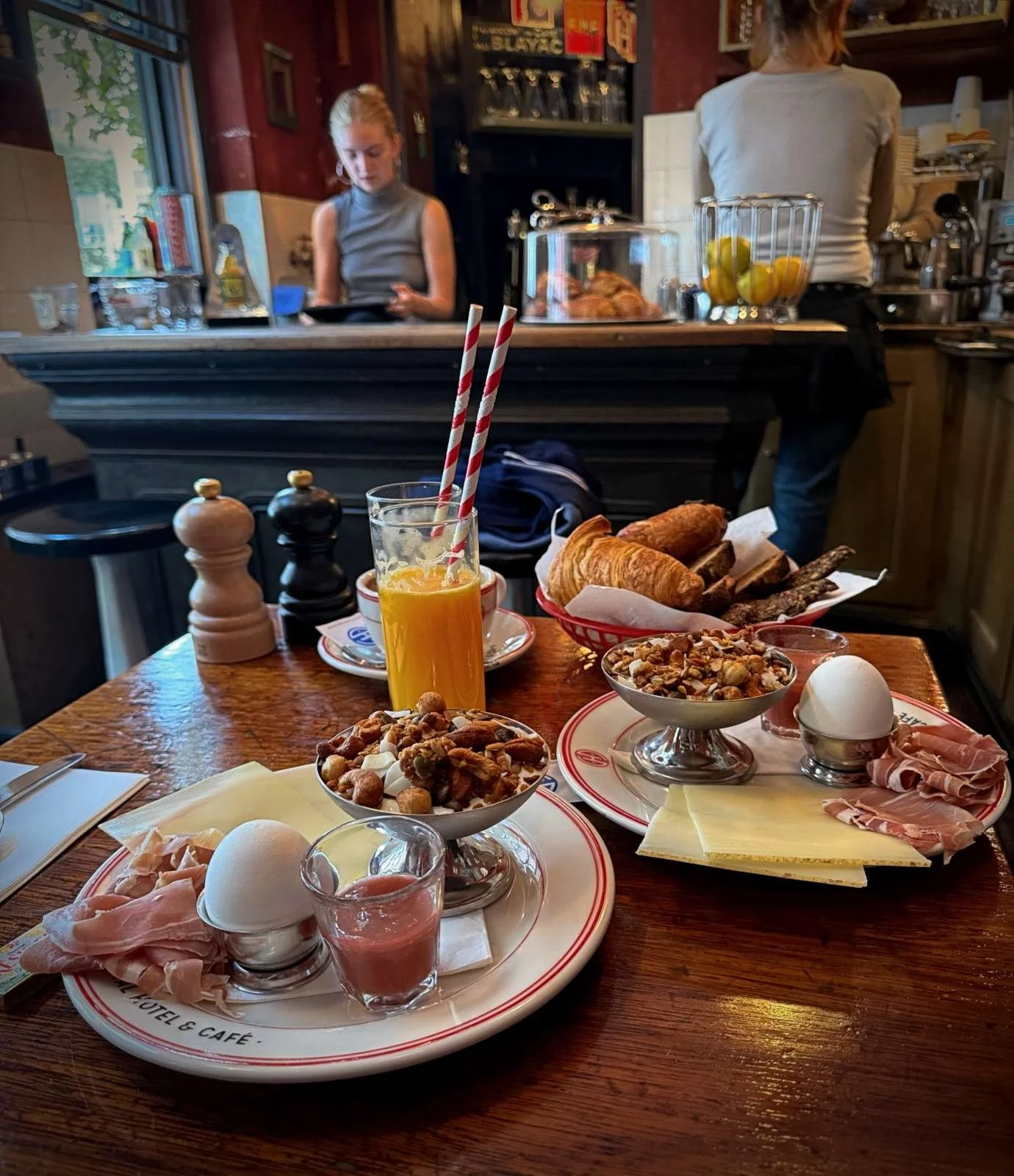 Ein gedeckter Frühstückstisch im Central Hotel & Café mit Orangensaft, Croissants, Müsli, gekochten Eiern, Schinken und Käse auf Tellern. Im Hintergrund ist eine Kaffeetheke mit einer Mitarbeiterin zu sehen.