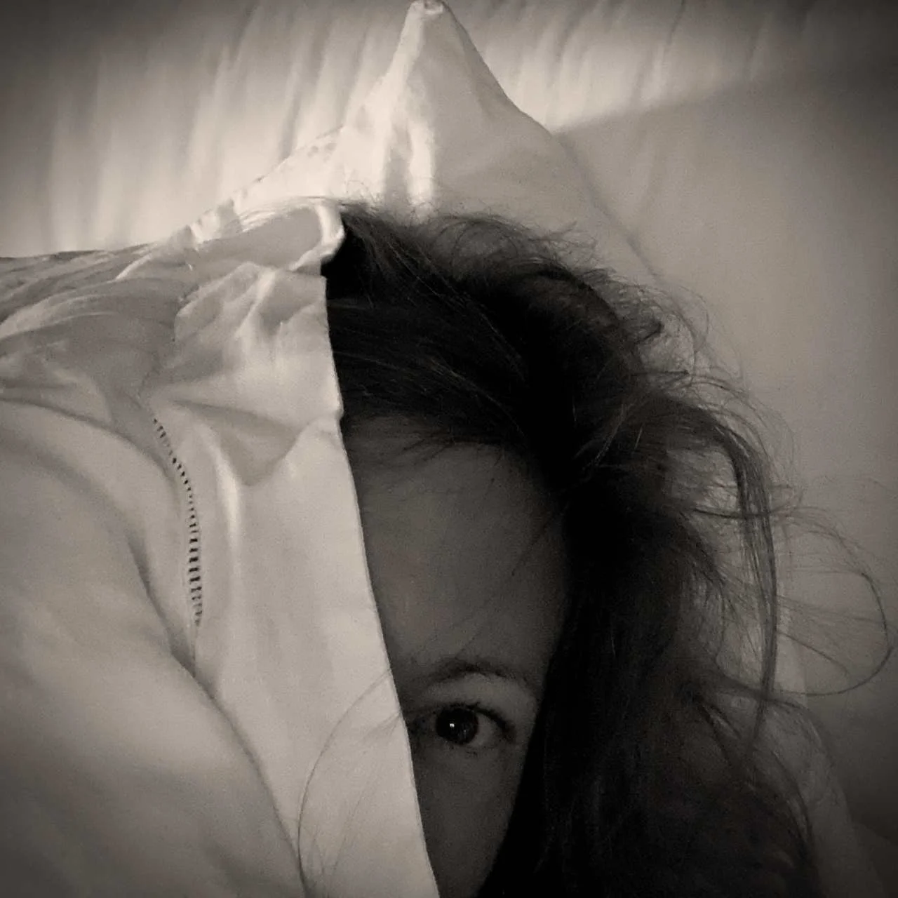 Schwarzweißaufnahme eines Gesichts, das halb unter einer weißen Bettdecke hervorschaut, mit unordentlichen Haaren auf einem Kissen.