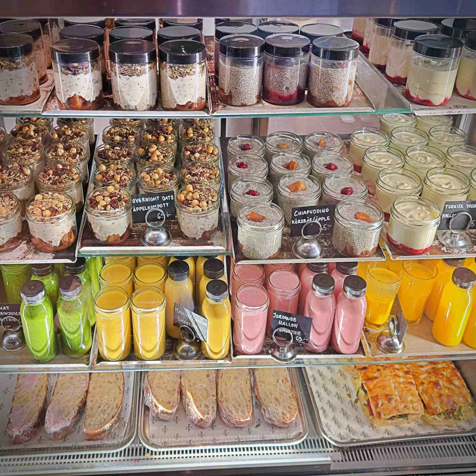 A well-stocked refrigerated display case filled with breakfast jars containing overnight oats and rhubarb chia pudding. Colorful bottled smoothies and fresh sandwiches are arranged on the shelves below.