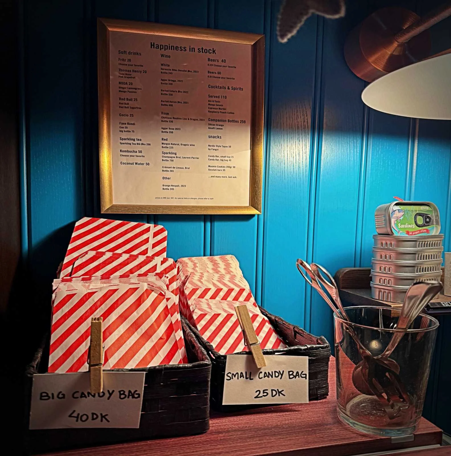 Im 25hours Hotel Paper Island Kopenhagen stehen rot-weiß gestreifte Papiertüten für Süßigkeiten in Körben. Ein Schild an der blauen Wand listet Getränke und Snacks unter dem Titel Happiness in stock.