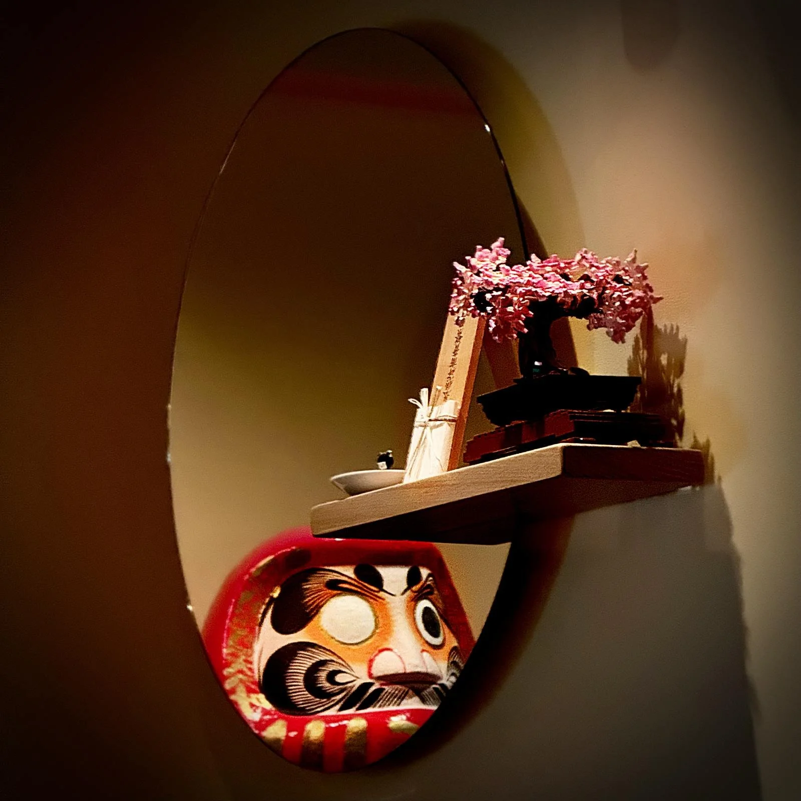 An oval mirror at Miyakodori restaurant in Stockholm reflects a red Daruma figure next to a shelf with a pink artificial bonsai tree.