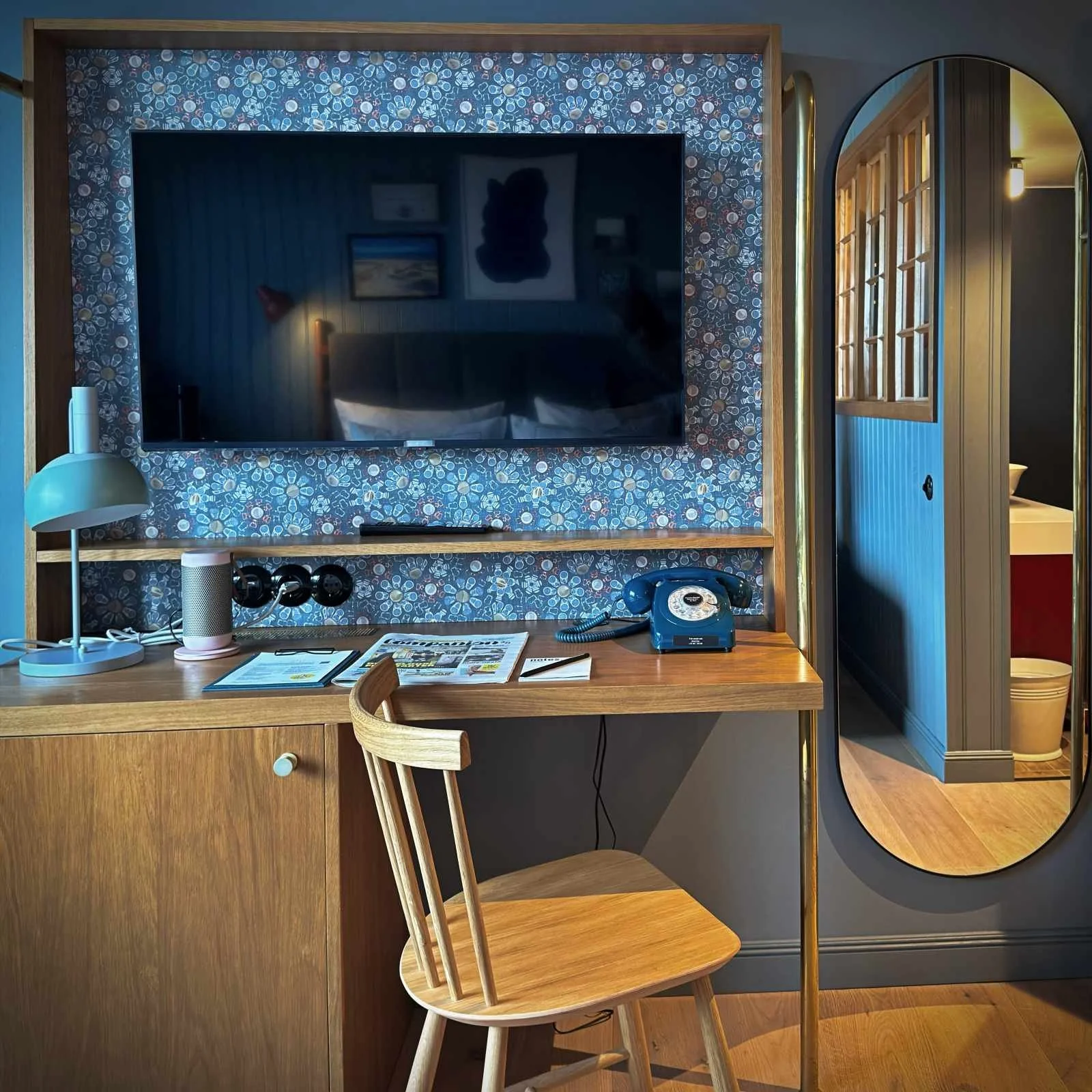 Arbeitsbereich im 25hours Hotel Paper Island Kopenhagen mit einem Holzschreibtisch, einem Stuhl davor und einem runden Spiegel an der blauen Wand. Auf dem Tisch stehen ein Telefon, eine Tasse und eine Glaskaraffe.
