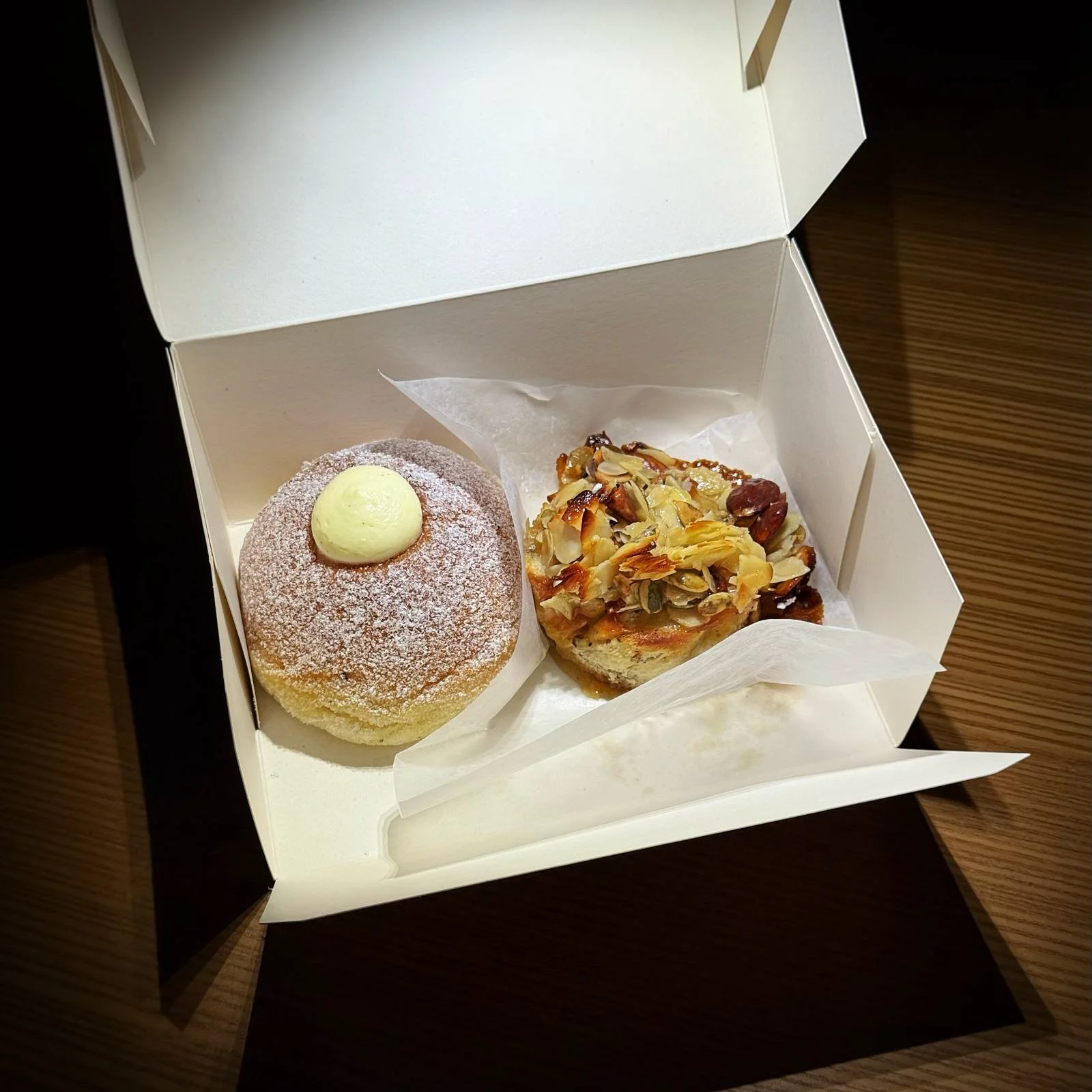 View inside an open white cardboard box containing two pastries. On the left is a cream-filled bun with powdered sugar, on the right a pastry topped with almonds.
