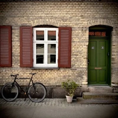 Black bicycle leaning against a light brick wall with a green door and shutters. The photo captures the characteristic Danish cityscape off the beaten track.