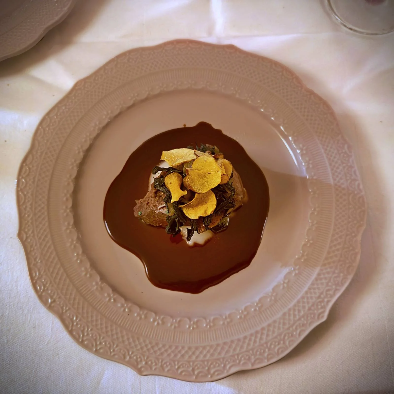 Ein beiger Teller mit Kalbsbacken in dunkler Rotweinsauce. Obenauf liegen gelbe Artischockenchips und grünes Gemüse. Es ist ein Hauptgang im Restaurant R21 in Amsterdam auf weißem Tischtuch.