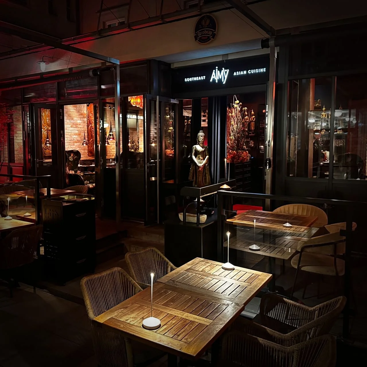 Abendliche Außenansicht des Aimy Restaurants mit überdachter Terrasse, beleuchteten Tischen, asiatischer Skulptur und Blick ins dekorierte Innere.