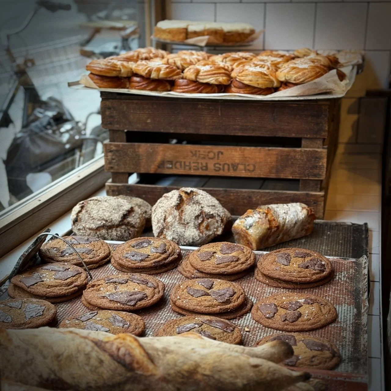 Large chocolate cookies fill the tray in the shop window. Behind them, sourdough loaves and fresh cinnamon buns lie on a wooden crate.