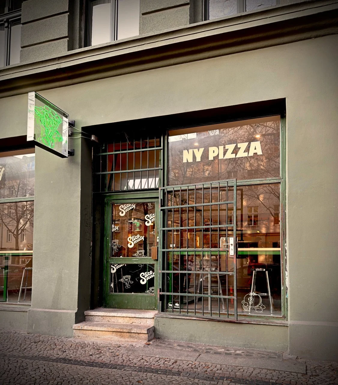 Exterior of Slice Society in Berlin featuring a green neon sign, metal gate and NY Pizza lettering on the window – view into the bar stools and interior.