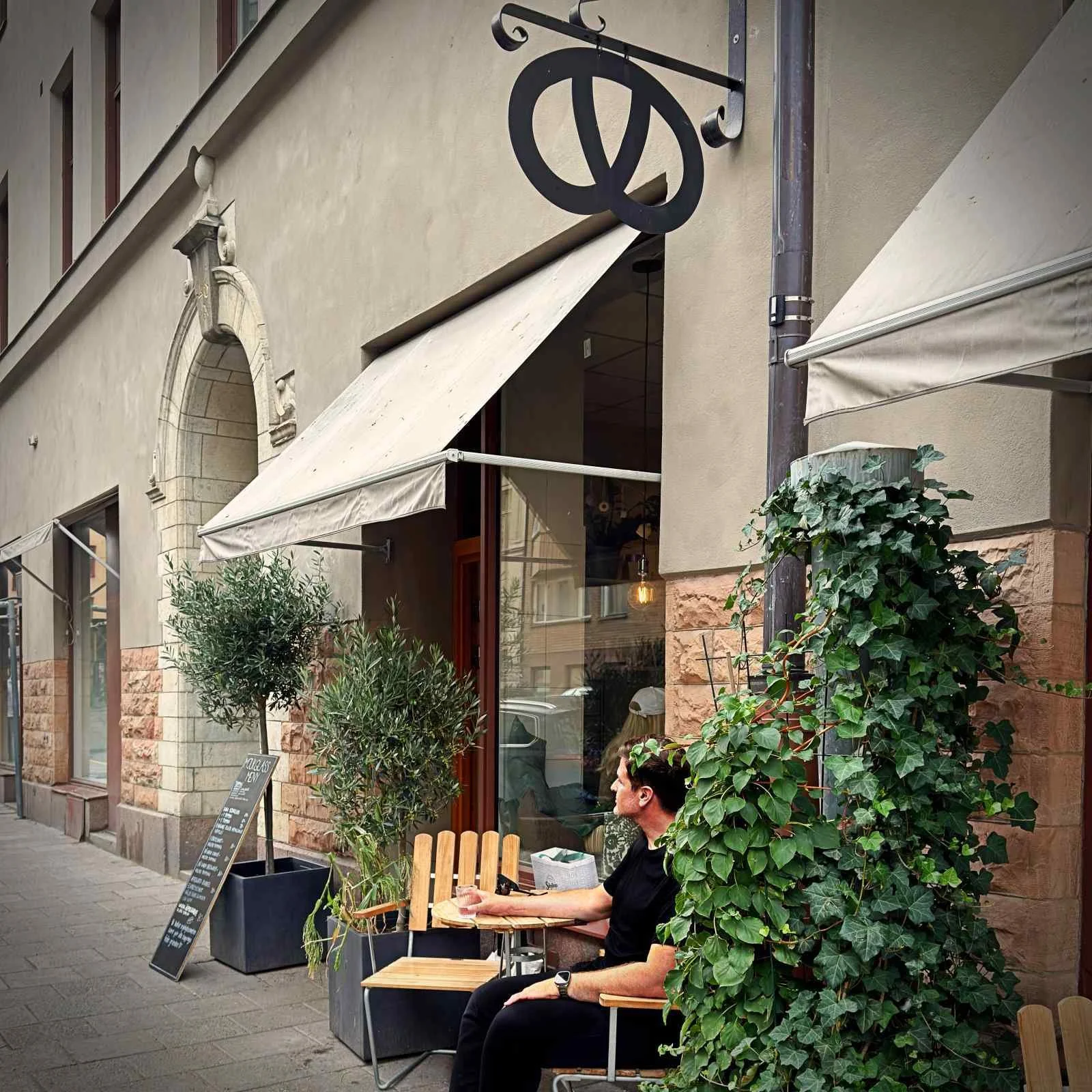 Eine Person sitzt an einem Metalltisch auf der Außenterrasse der Bergstrands Bageri in Stockholm; auf dem Tisch stehen Kaffeetassen und ein Teller mit Gebäck, im Hintergrund ist eine Straße sichtbar.