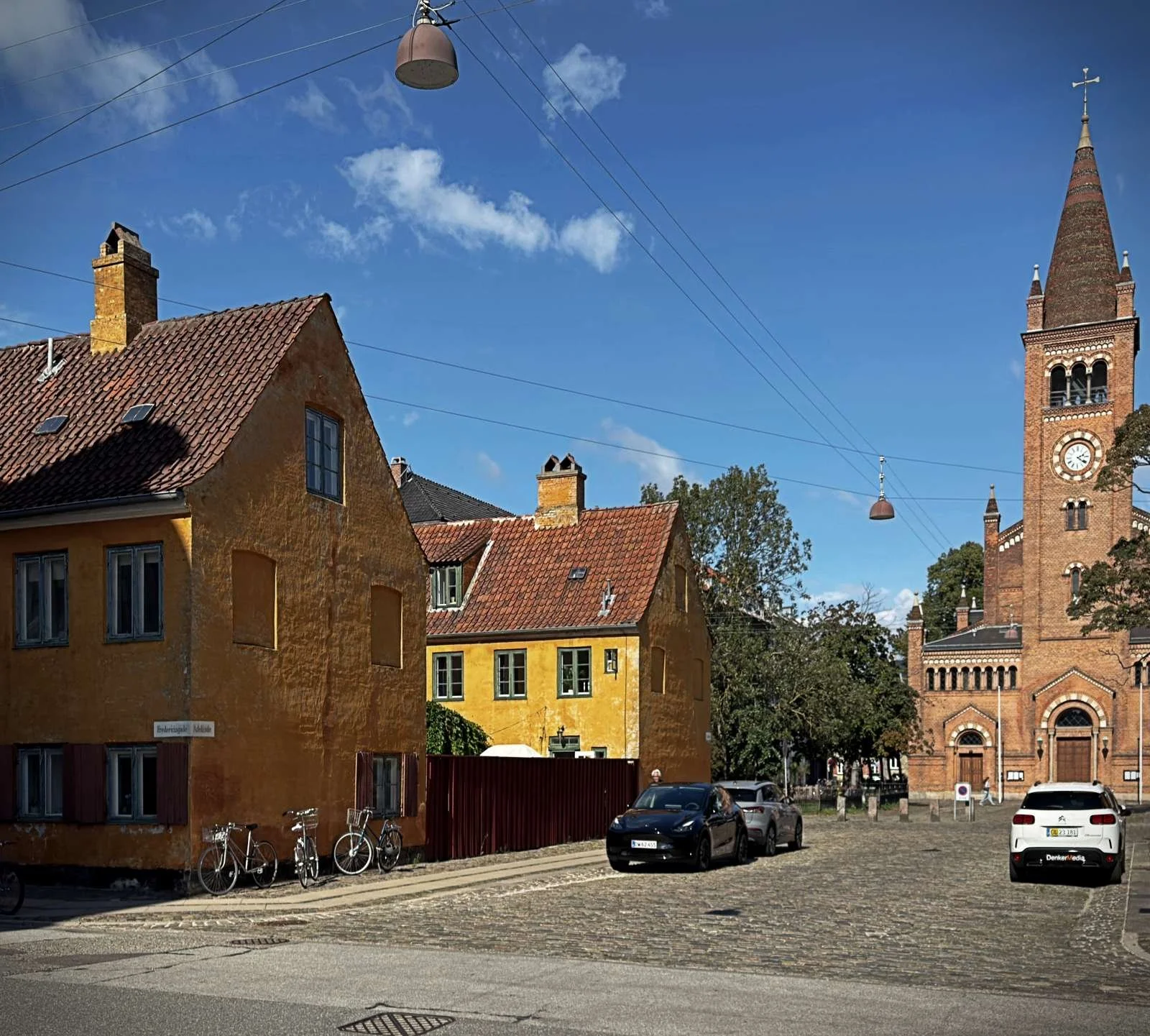 A cobbled square with yellow houses and a brick church in the Nyboder district of Copenhagen. Bicycles lean against the houses on the left, while cars are parked in front of the church with a clock tower on the right.