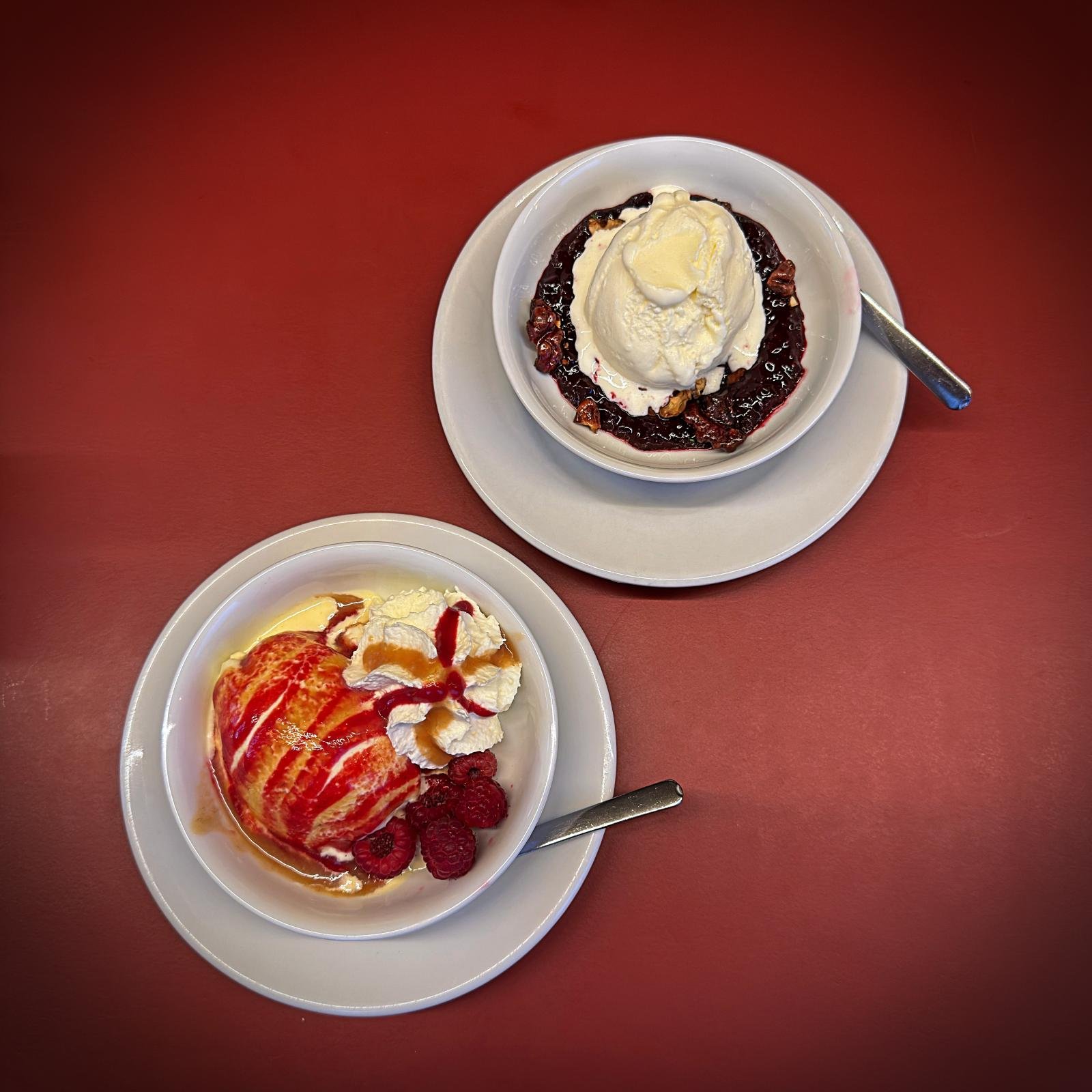 Top-down view of two bowls with ice cream desserts on a red table. One bowl contains ice cream with whipped cream and raspberries, the other ice cream on berry compote.