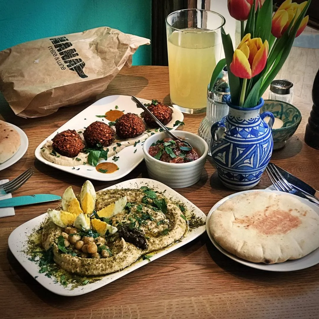 Wooden table at restaurant Nana Meze & Wine with hummus, egg, falafel, pita bread, a bowl of dip, and tulips in a ceramic vase.