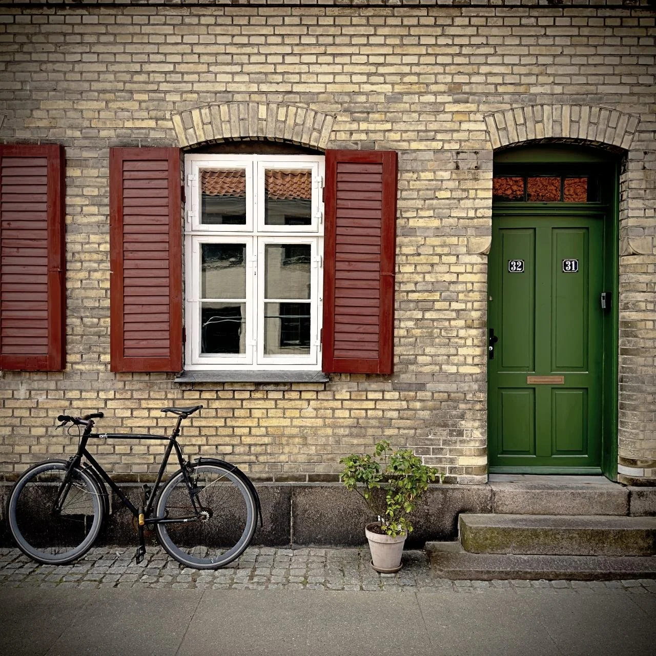 Historic brick house in Nyboder, Copenhagen, featuring a yellow facade, white-framed windows and red wooden shutters. Green front door, bicycle against the wall, small potted plant on cobblestones.