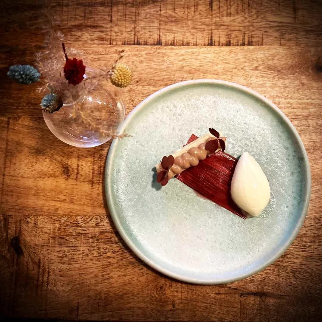 Ein Dessert aus Rhabarber, Milchschokolade und einer Nocke Eis auf einem hellblauen Keramikteller im Restaurant Selma in Kopenhagen. Daneben steht eine kleine Vase mit Trockenblumen auf dem Holztisch.