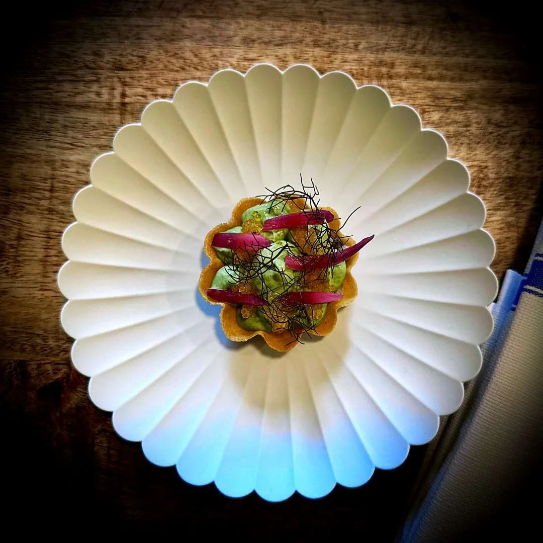 Green anise herring with nettle, roe, and fennel served on a white fluted plate at Restaurant Selma in Copenhagen. The dish is placed on a wooden table next to a striped napkin.