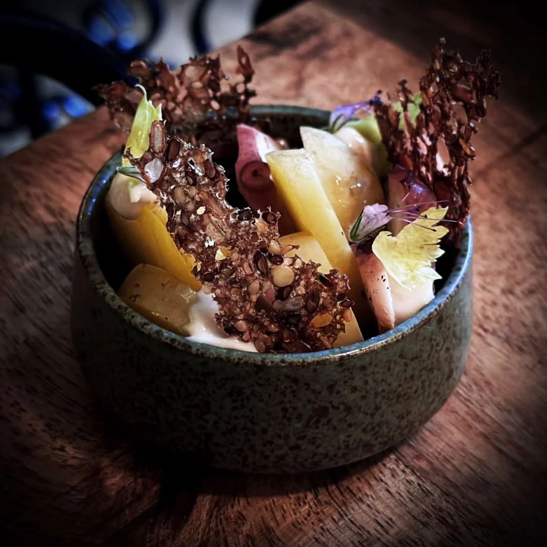 Eine Schale mit Pulpo, neuen dänischen Kartoffeln, Sellerie und Minze im Restaurant Selma in Kopenhagen. Das Gericht wird mit knusprigen Saatchips serviert und steht auf einem dunklen Holztisch.