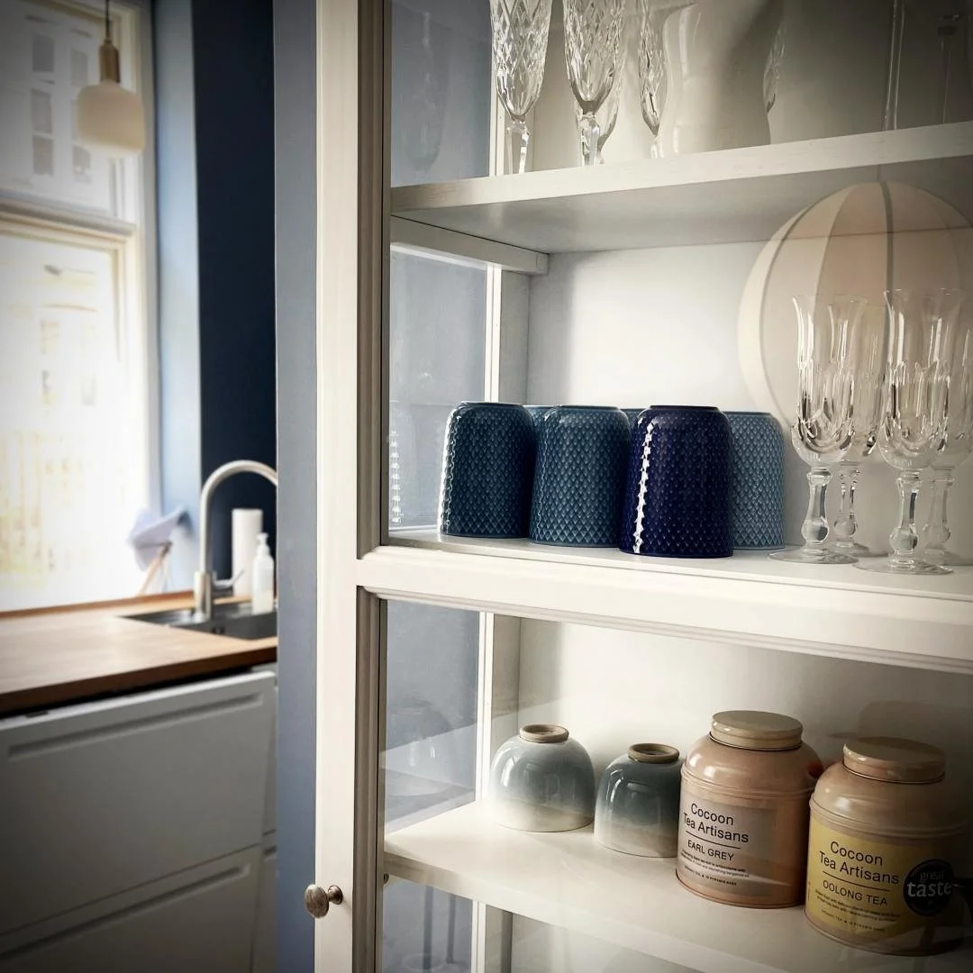 View into the kitchen of an Airbnb on Haregade in Copenhagen. In the foreground is a white glass cabinet displaying crystal glasses, blue mugs, and tea canisters from the brand "Cocoon Tea Artisans"